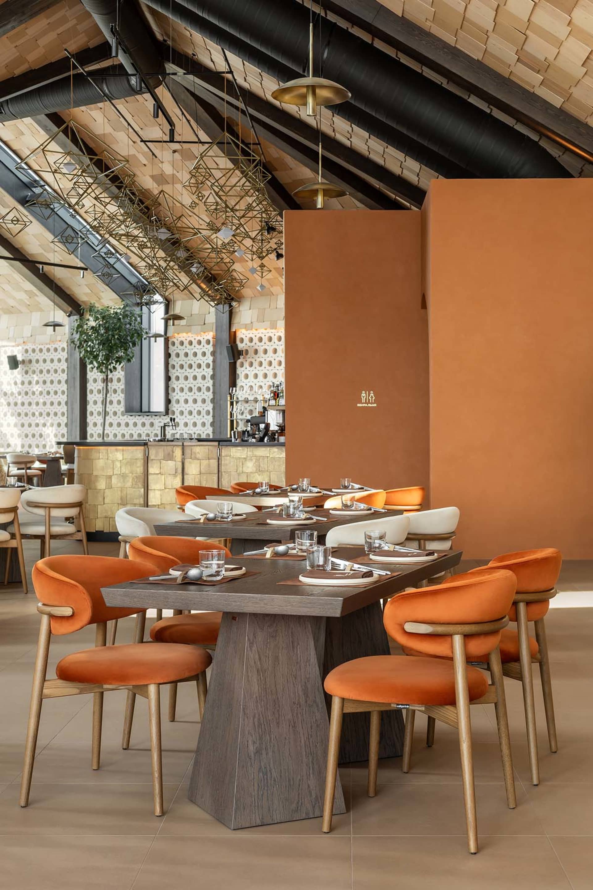 Modern restaurant interior with dark wood dining tables, burnt orange velvet upholstered chairs, geometric brass pendant lights, and a vaulted wood ceiling with exposed black ducts.