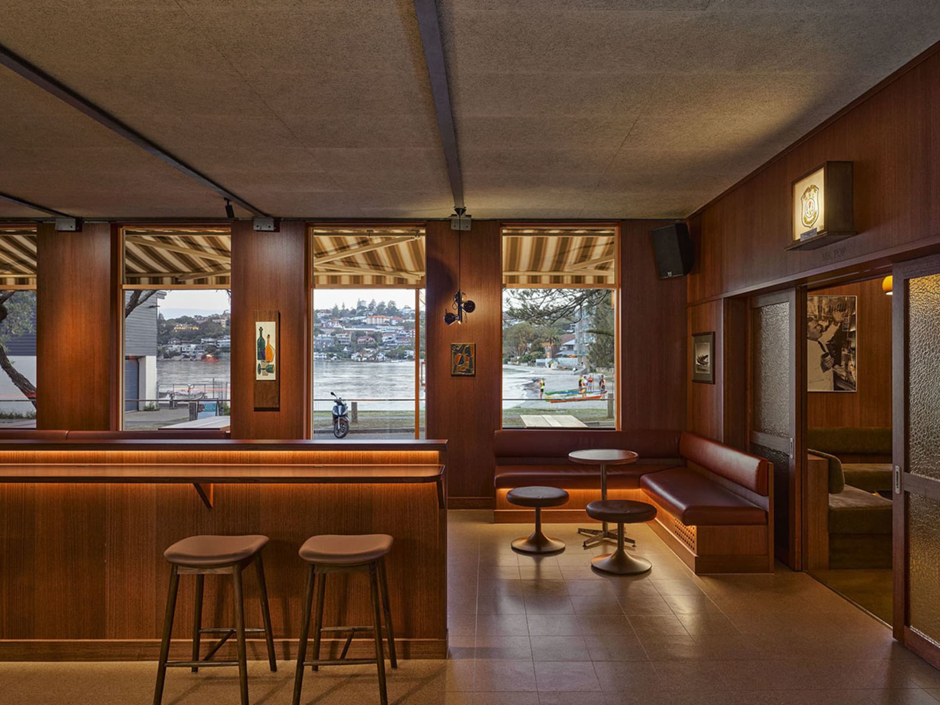 Mid-century modern cafe interior featuring a long teak wood bar, leather banquette seating, round pedestal tables, and large windows overlooking a harbor with boats and distant hills.