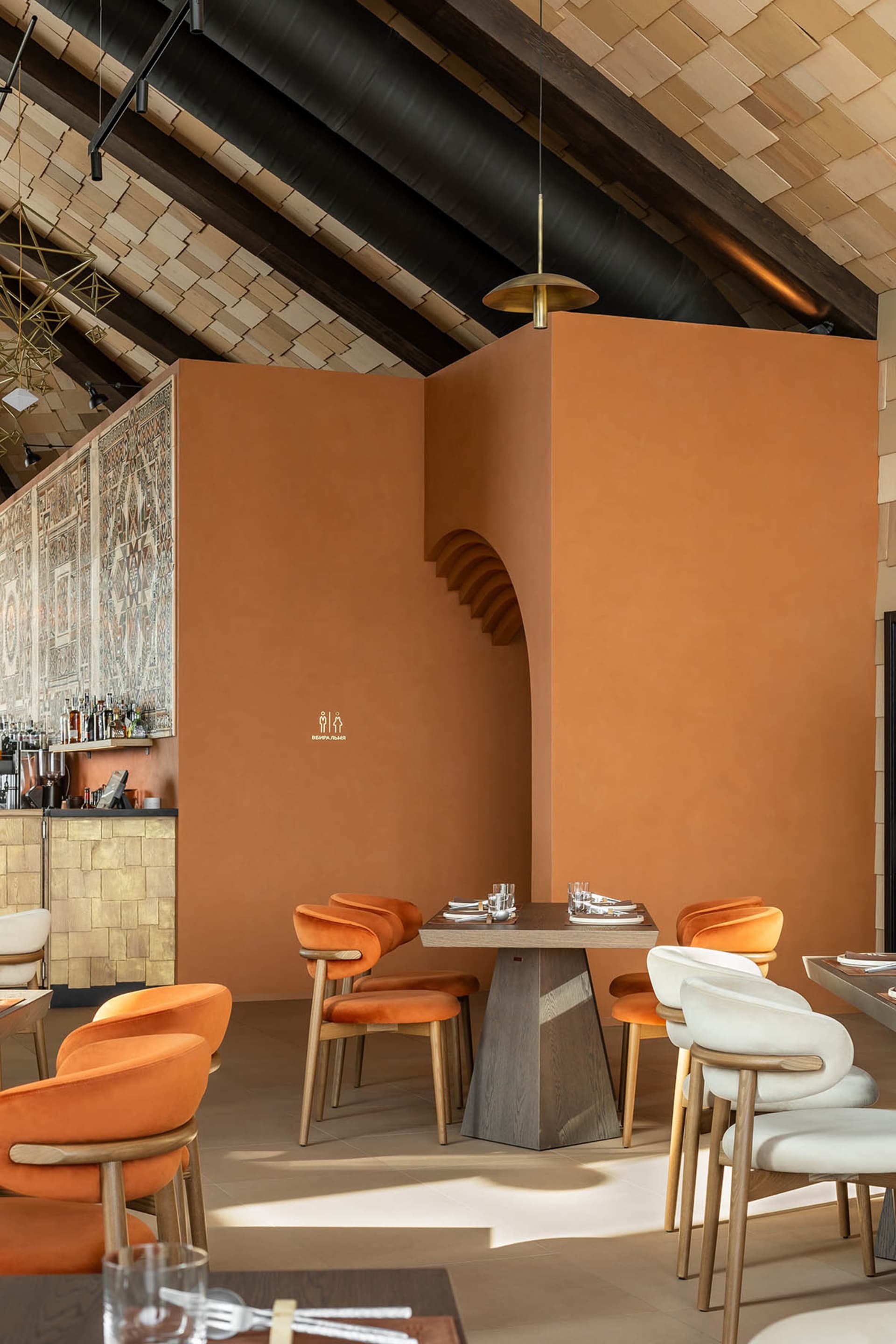 Modern restaurant interior with terracotta plaster walls, burnt orange and cream velvet dining chairs, wooden tables, and a striking high ceiling with exposed dark beams and wooden slats.
