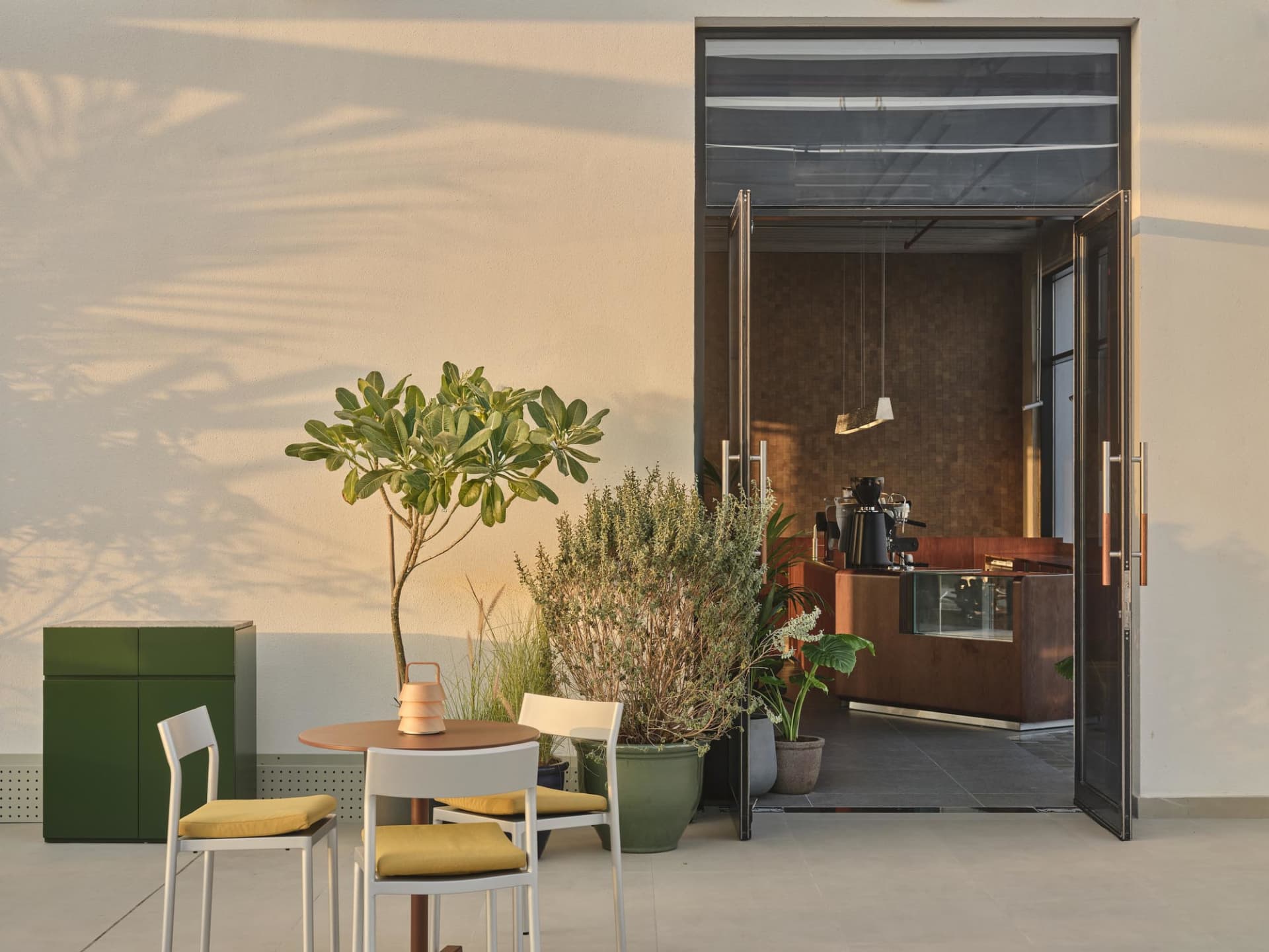 Modern outdoor cafe patio featuring a round wooden table with white chairs and yellow cushions, surrounded by lush potted plants, a green storage cabinet, and an open view into a wood-paneled coffee station with hanging light fixtures.