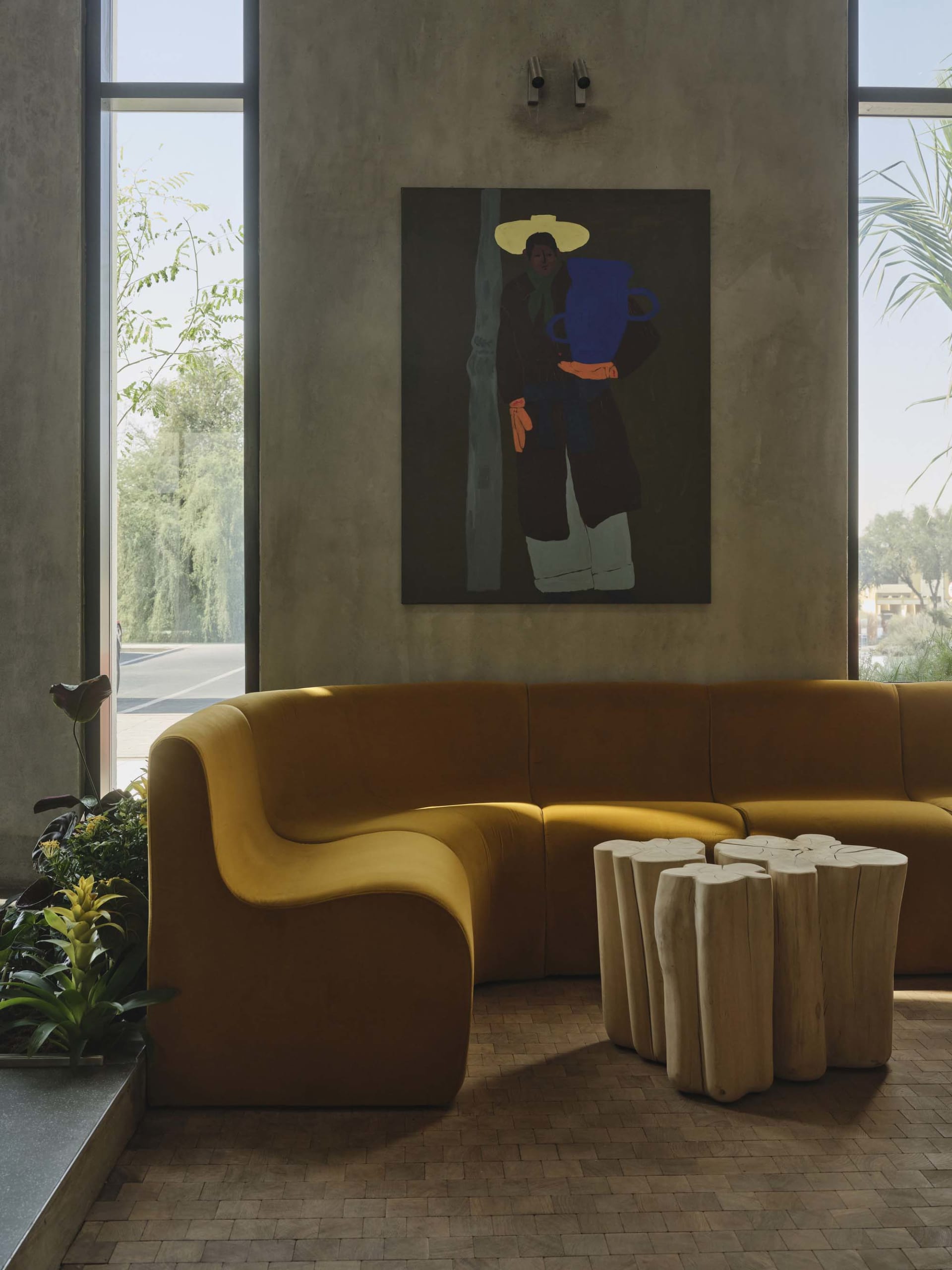 Modern living room featuring a curved mustard-yellow velvet sofa, two light wood stump coffee tables, a large colorful painting of a figure, concrete walls, and large windows with natural light.