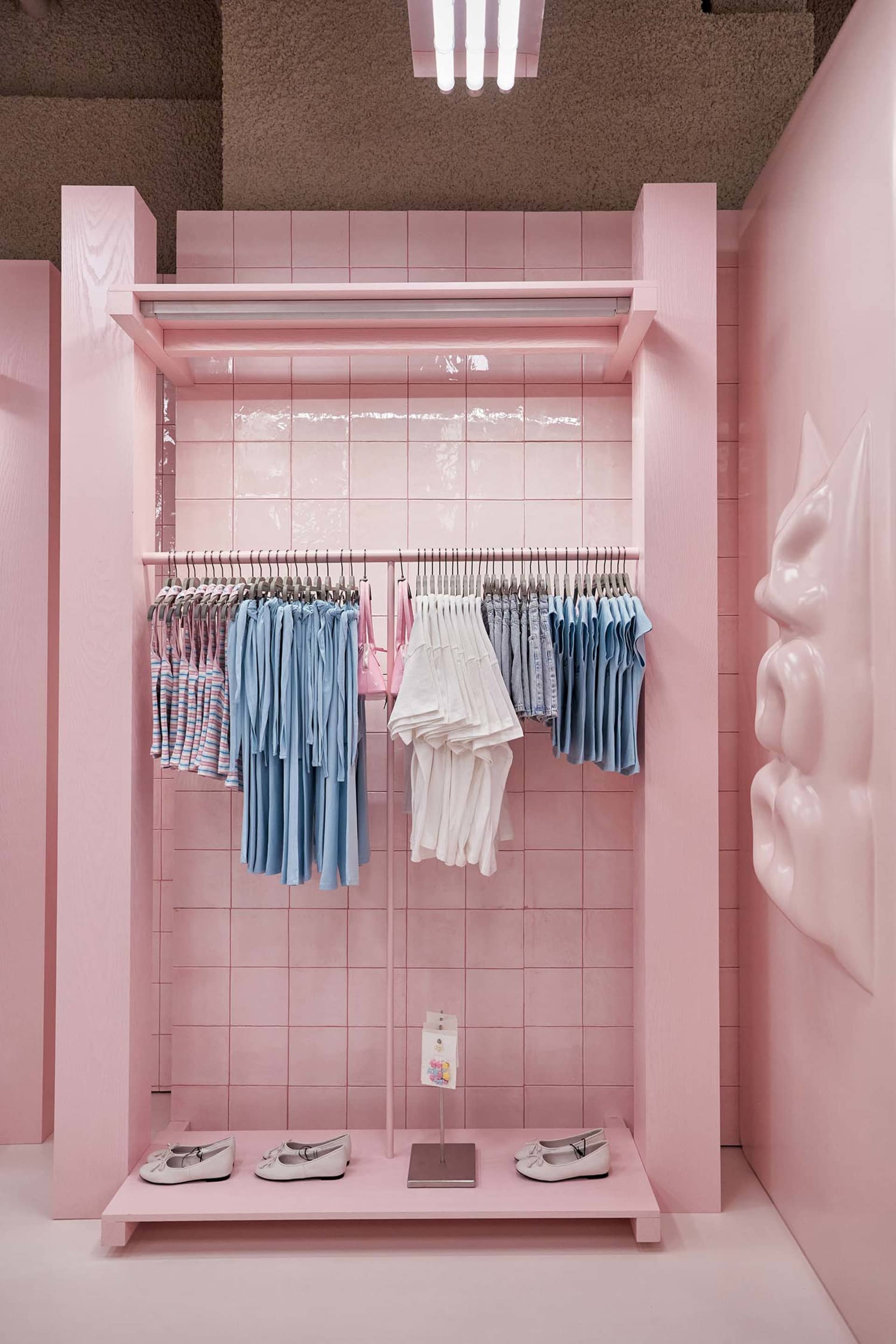Pink tiled retail display alcove with clothing racks holding pastel and striped garments, white ballet flats on a pink platform, and a large sculptural pink face on the right wall.