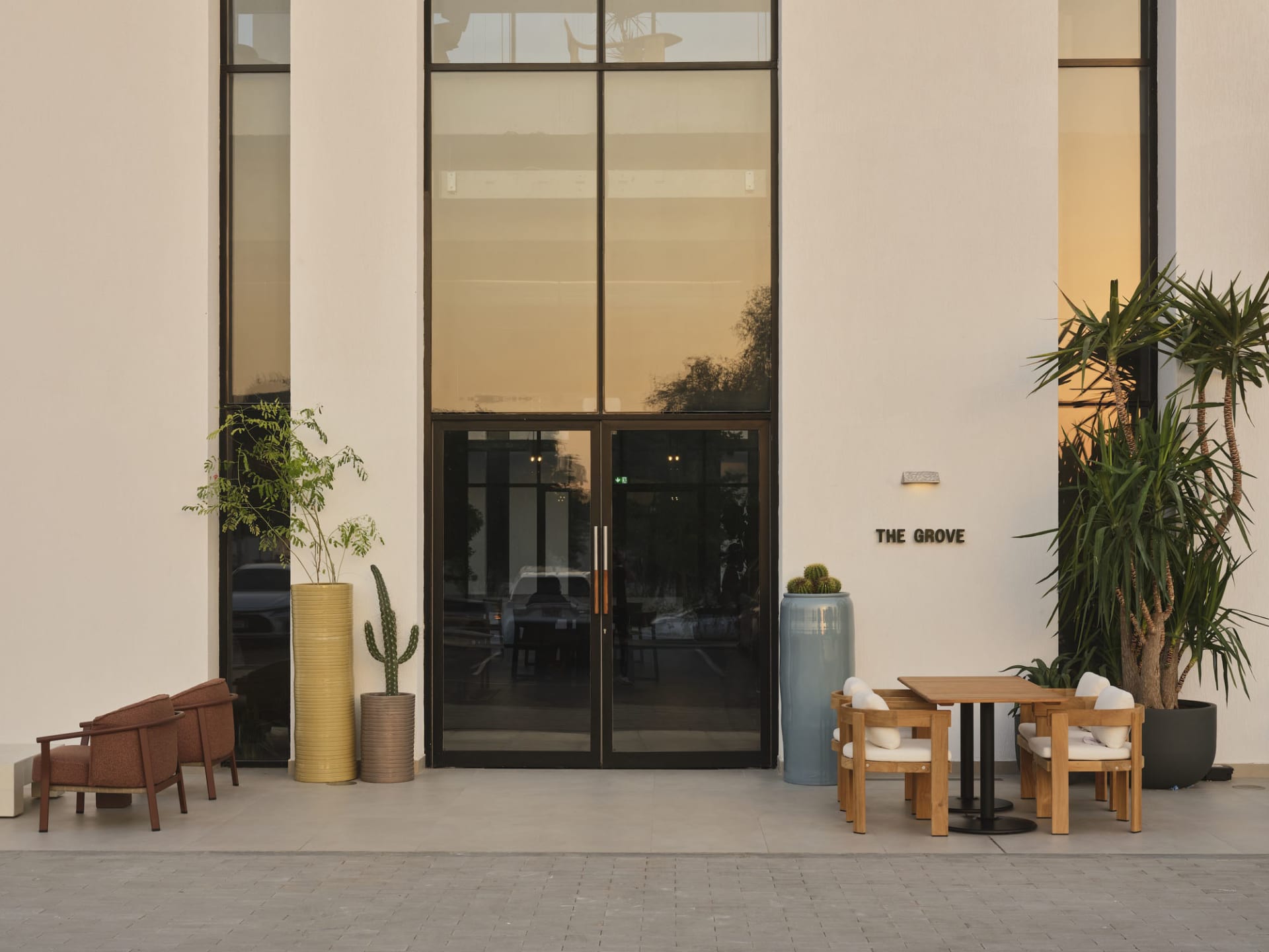Modern minimalist building entrance with wooden armchairs, a square dining set, large colorful planters, and tall glass doors at dusk.