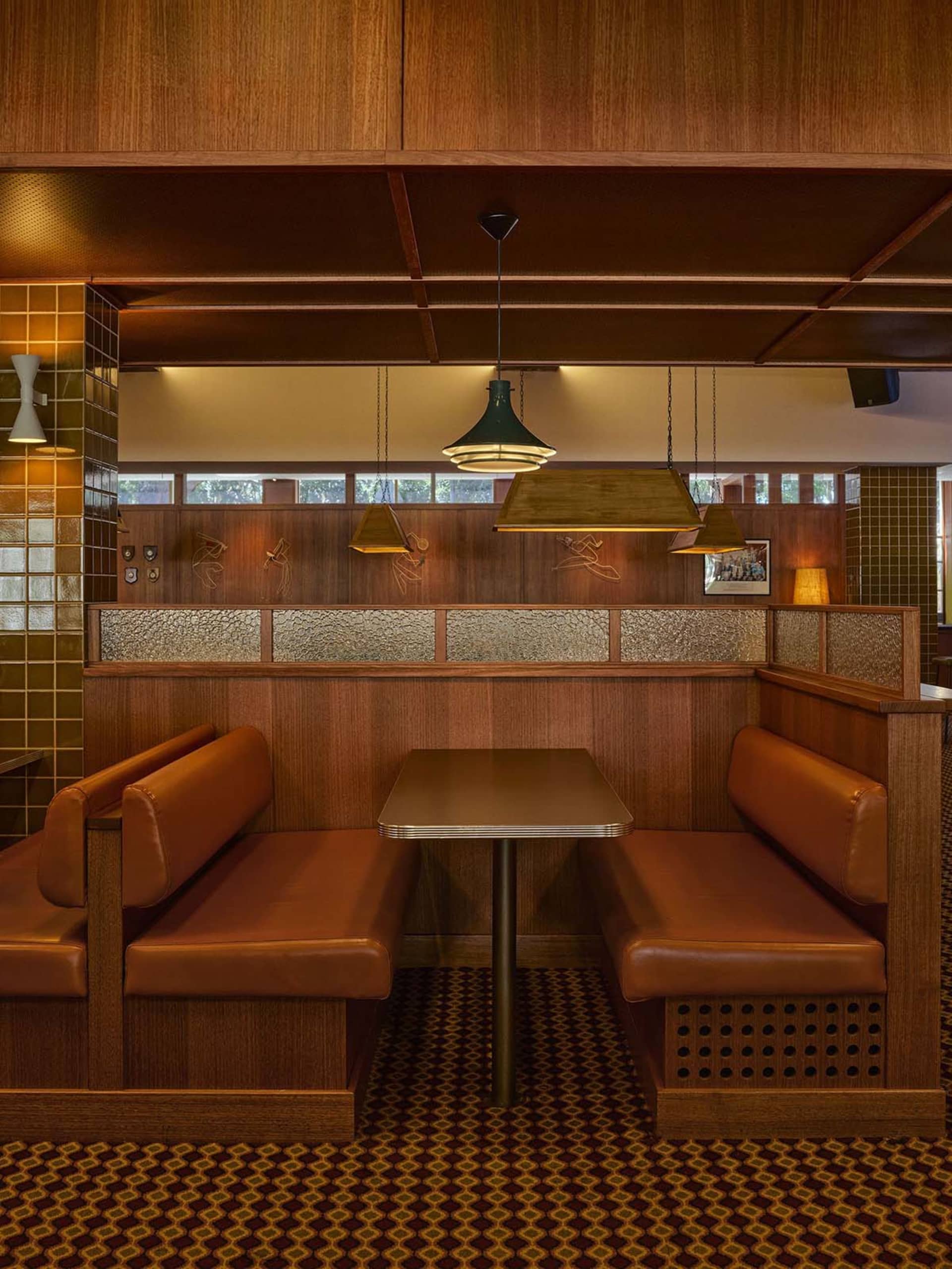 Vintage-style restaurant booth seating with brown leather upholstery, dark wood paneling, patterned carpet, and pendant lights in a warm retro interior.