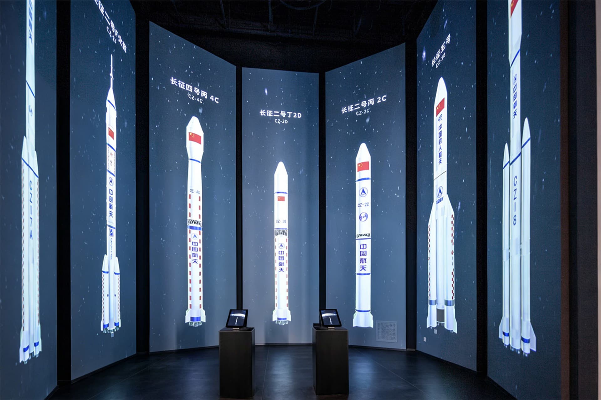 Dark exhibition room with curved walls covered in large LED screens displaying various Chinese Long March rockets against a starry background, with two interactive kiosks in the center.