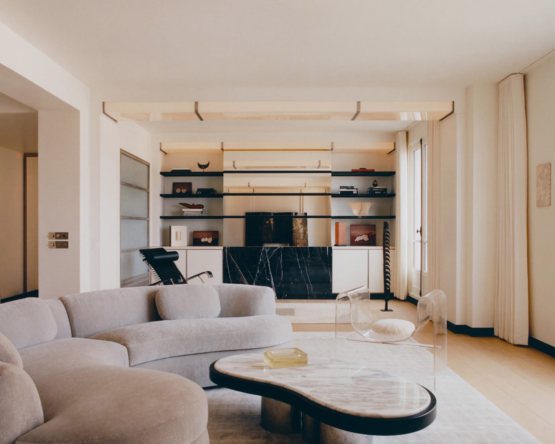 Modern minimalist living room with a curved black and white marble coffee table, light beige curved sectional sofa, transparent acrylic chairs, and built-in black floating shelves against a marble accent wall.