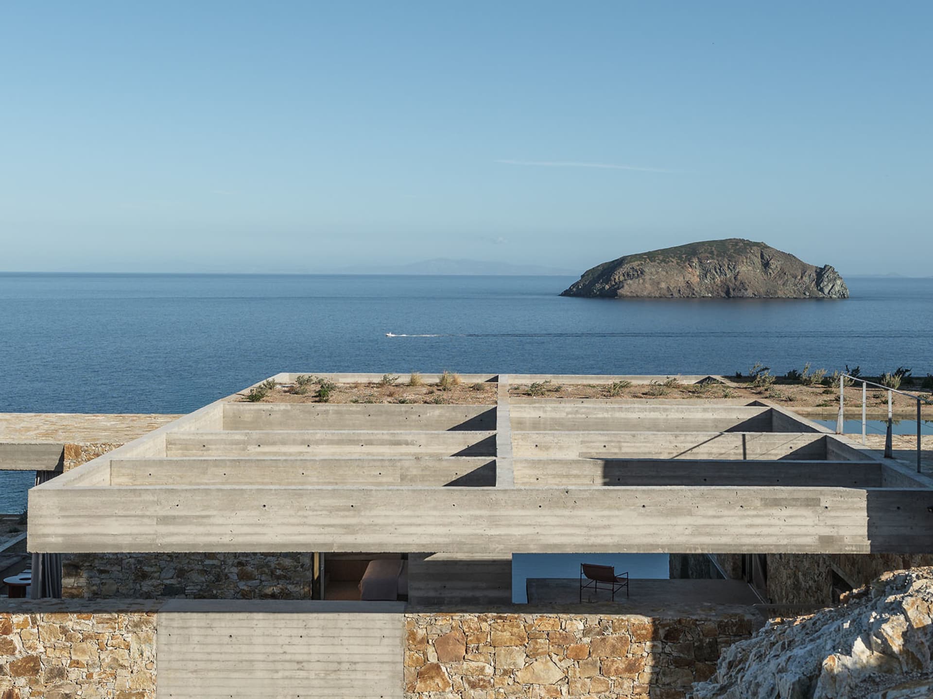 Modern exposed concrete rooftop terrace overlooking the deep blue Aegean Sea with a small boat and rocky island in the distance.