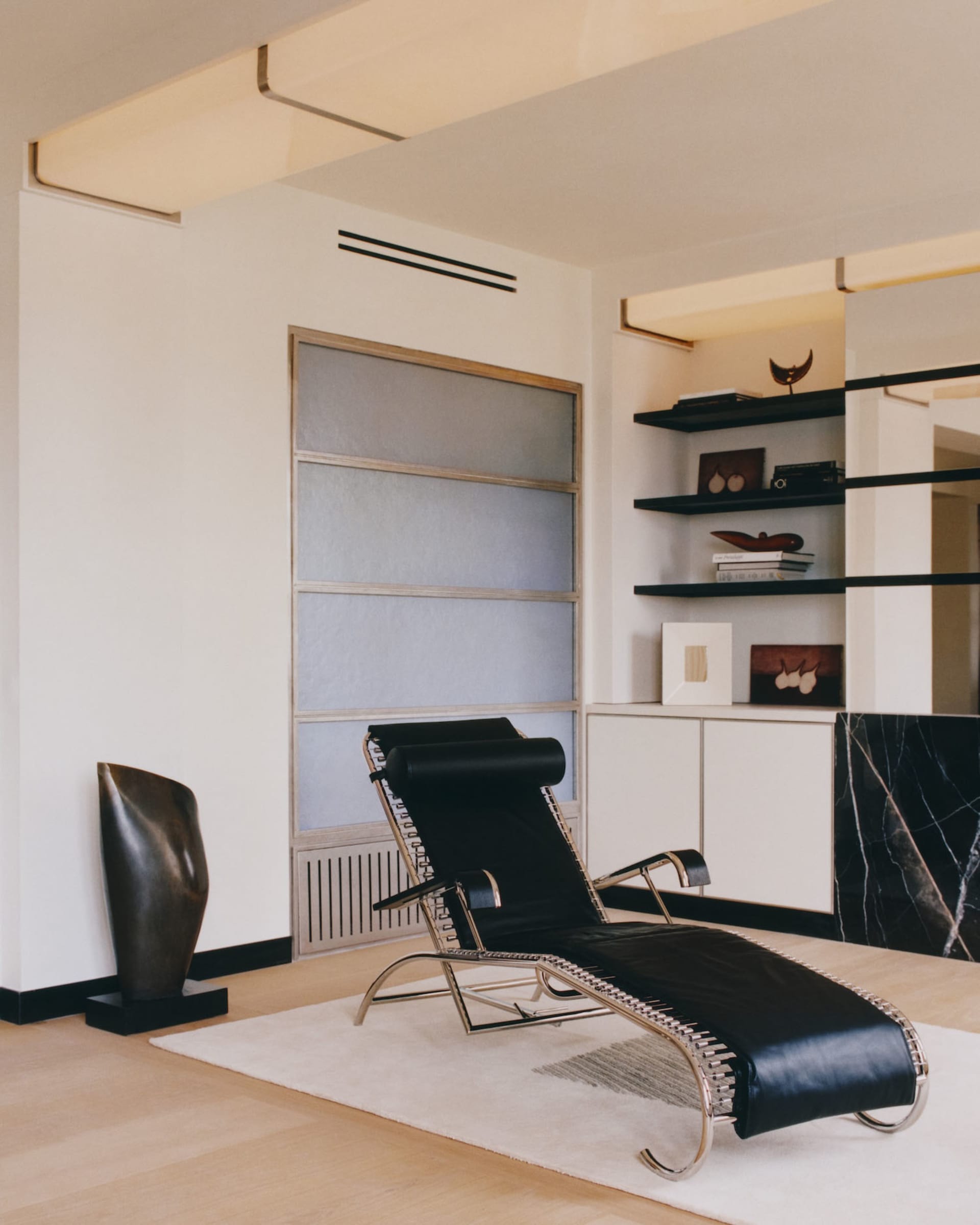 Modern minimalist living room featuring a black leather and chrome chaise lounge on a white rug, with built-in shelving, marble accent wall, and abstract bronze sculpture.