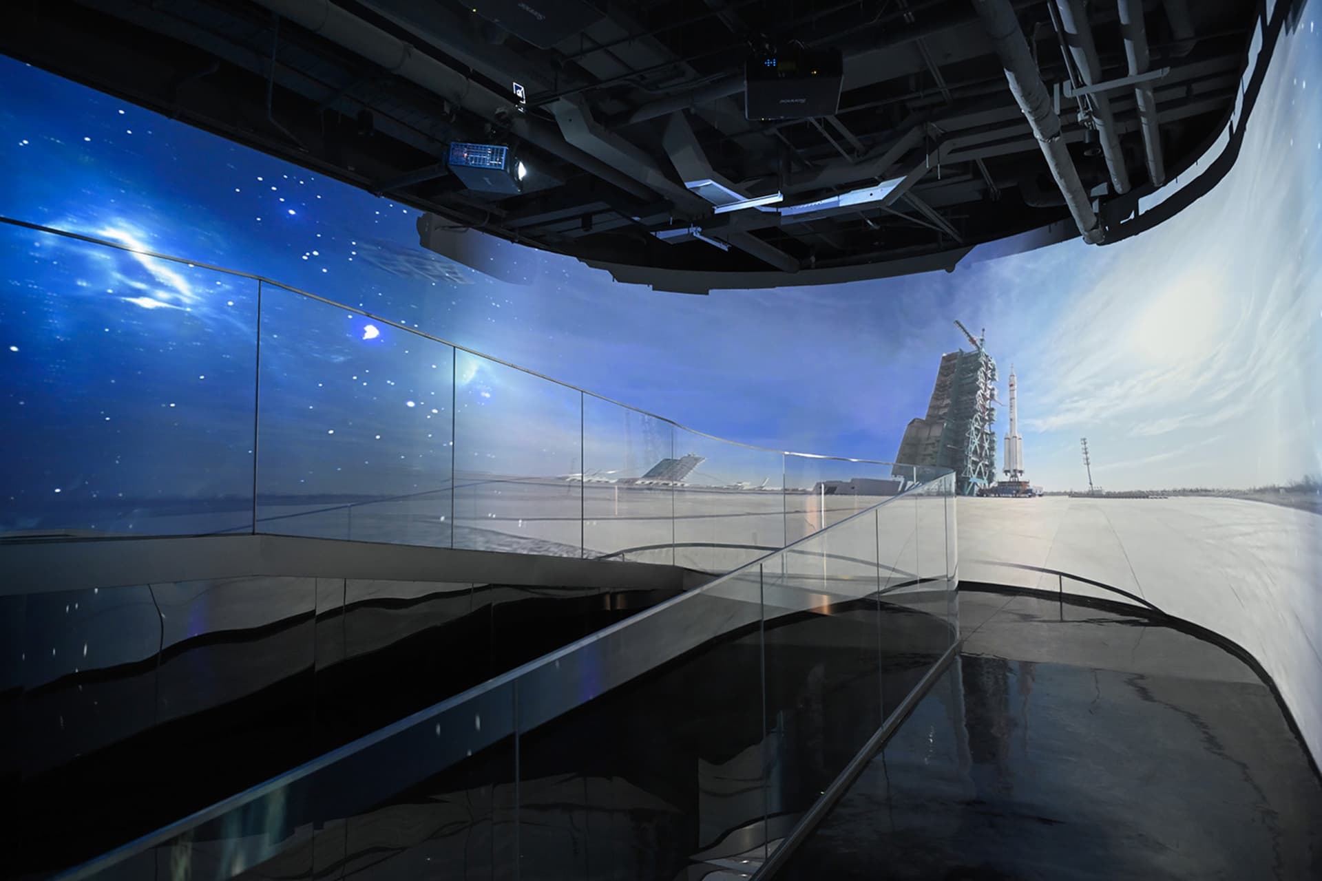 Curved glass and steel walkway inside the Kennedy Space Center with immersive panoramic projections of a starry nebula on one side and a rocket on the launch pad on the other.
