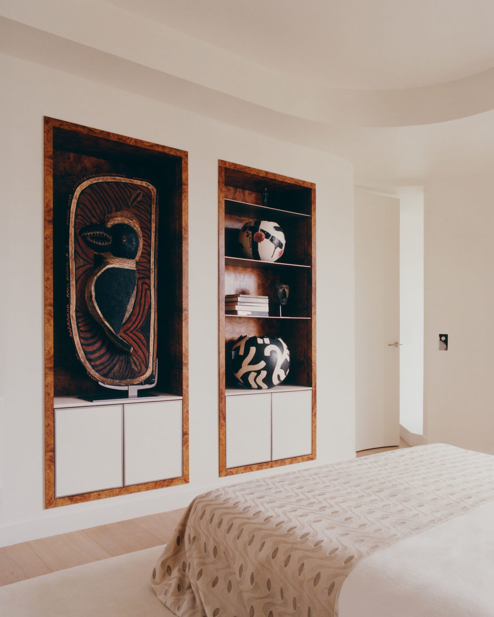 Modern bedroom featuring two recessed burl wood display niches filled with African tribal masks and pottery, white lacquered cabinetry below, and a bed with patterned beige throw in the foreground.