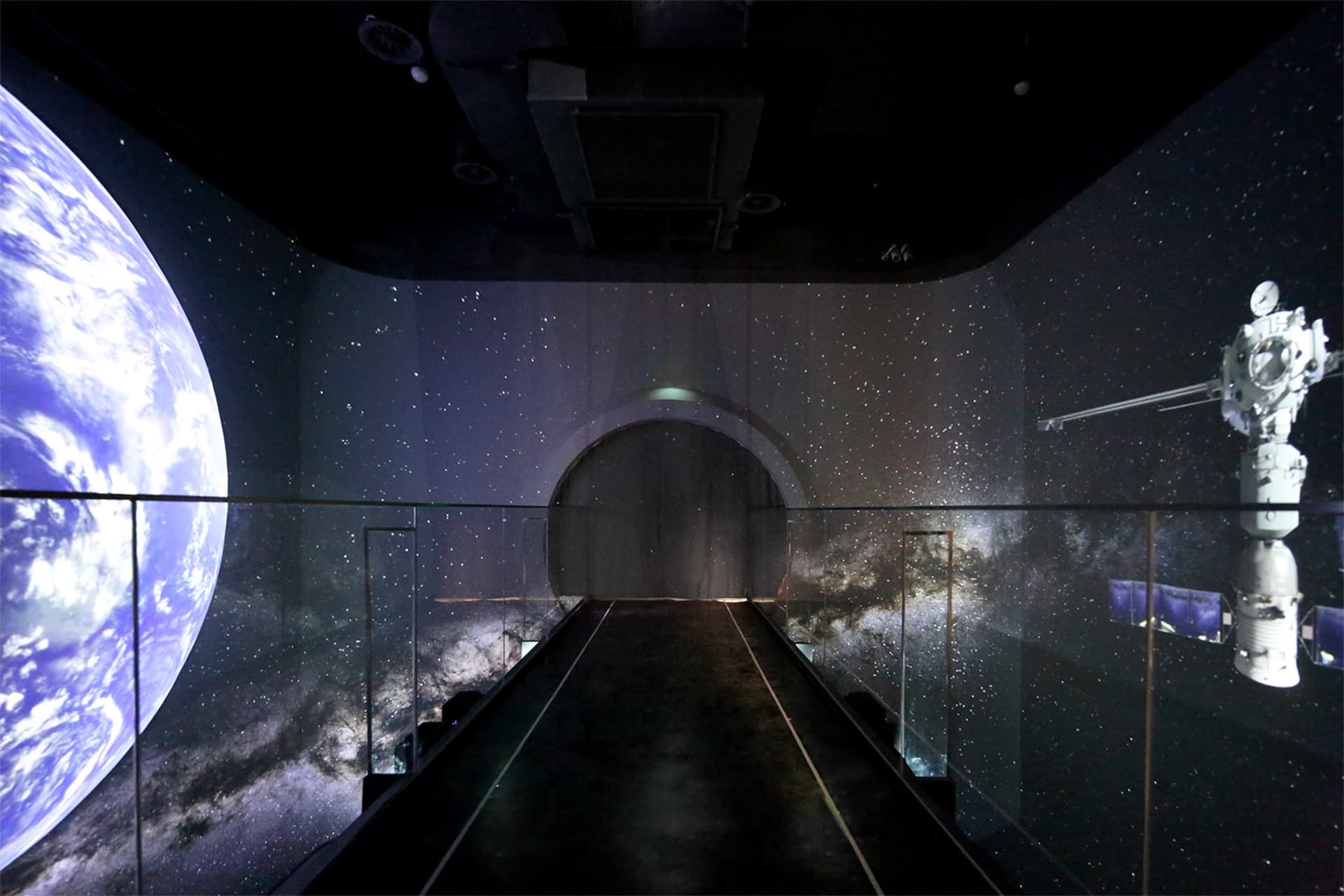 Dark immersive exhibition space featuring a central walkway with glass railings, surrounded by large-scale projections of planet Earth, the Milky Way, stars, and a detailed satellite model against a space backdrop.