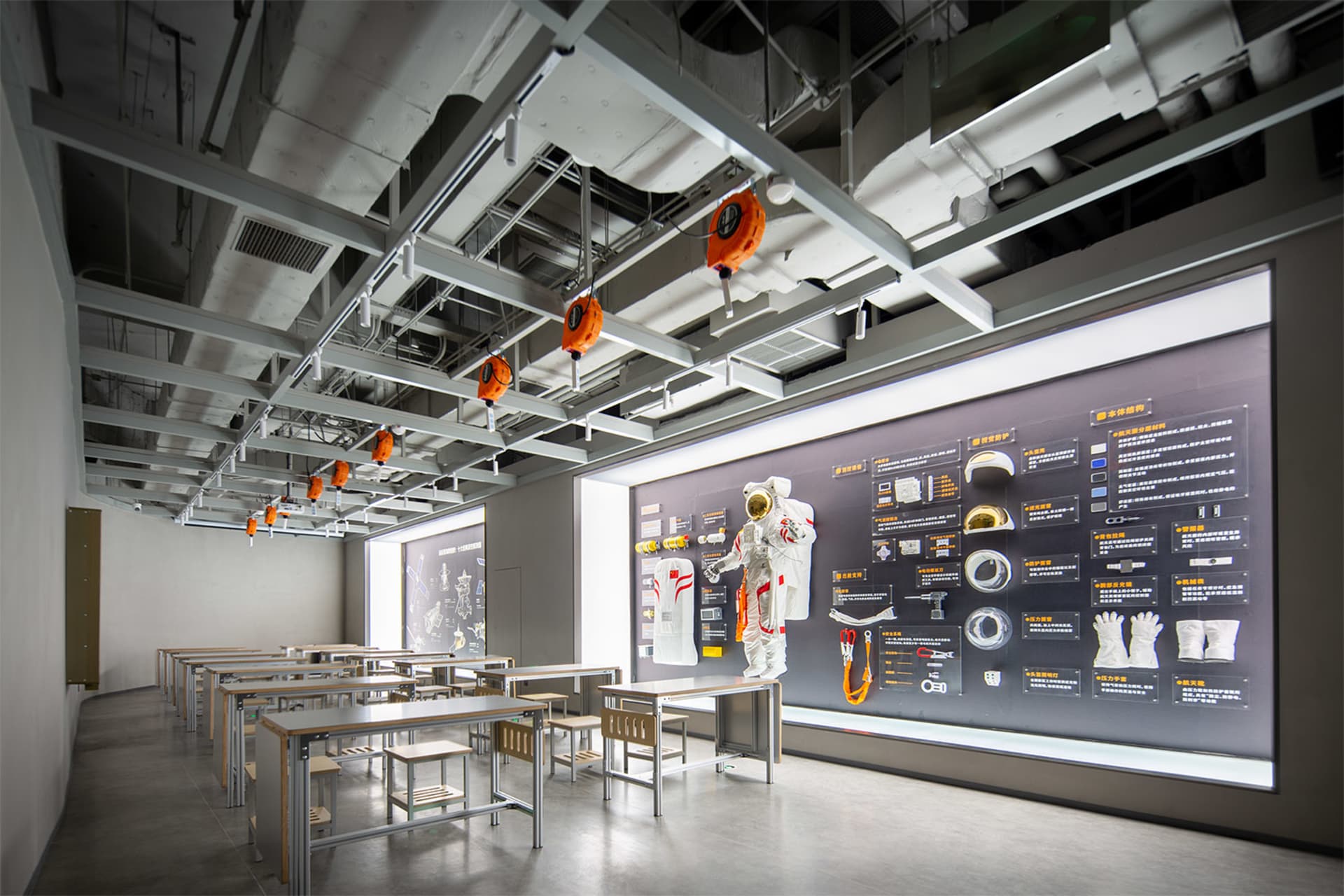 Modern classroom interior featuring rows of steel and wood desks, a full white-and-orange astronaut suit suspended from the ceiling, and a large illuminated interactive exhibition wall displaying aerospace equipment and diagrams.