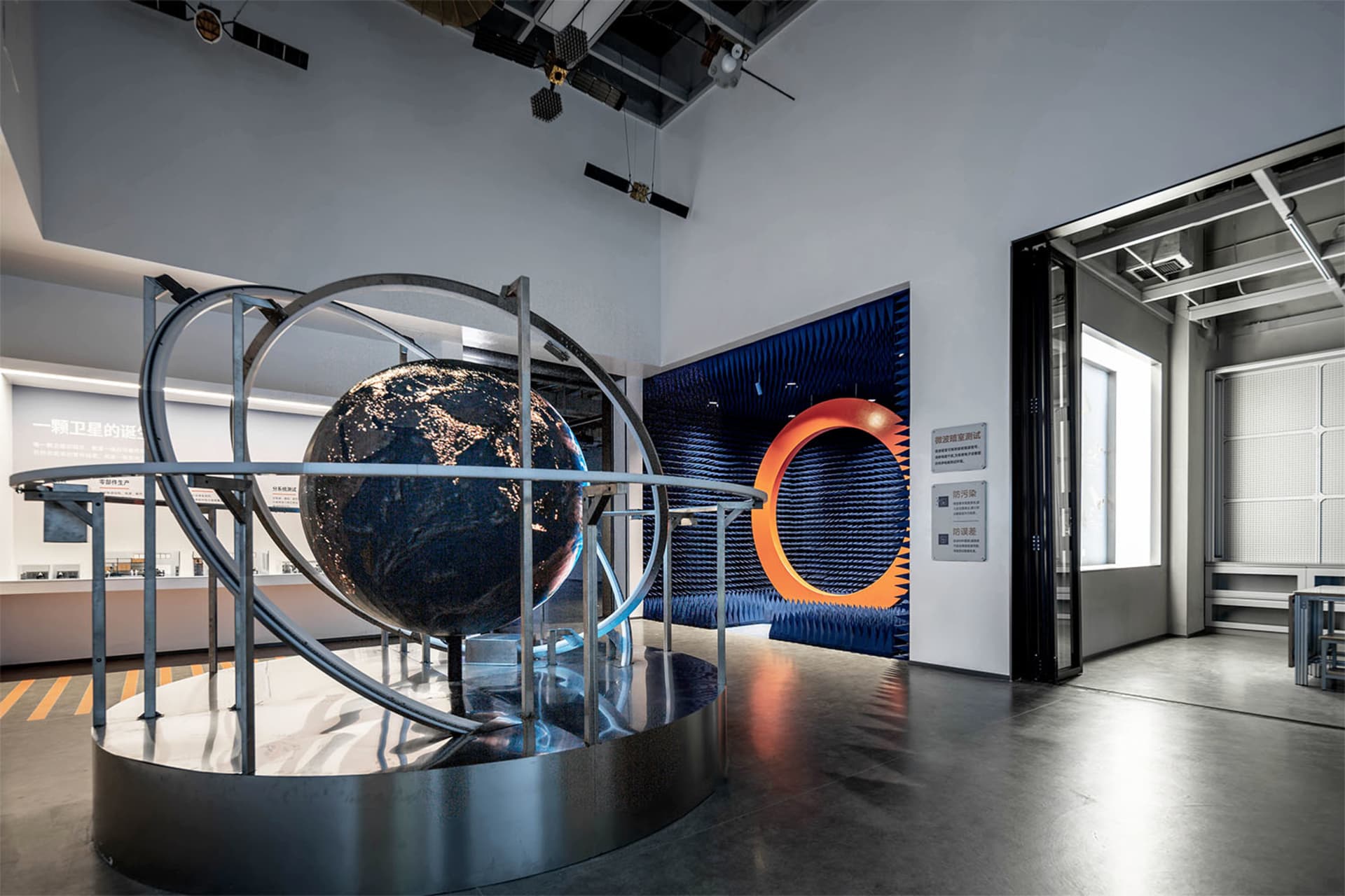 Large metallic globe exhibit with orbital rings displayed on a circular platform inside a contemporary science museum with blue acoustic panels and industrial ceiling.