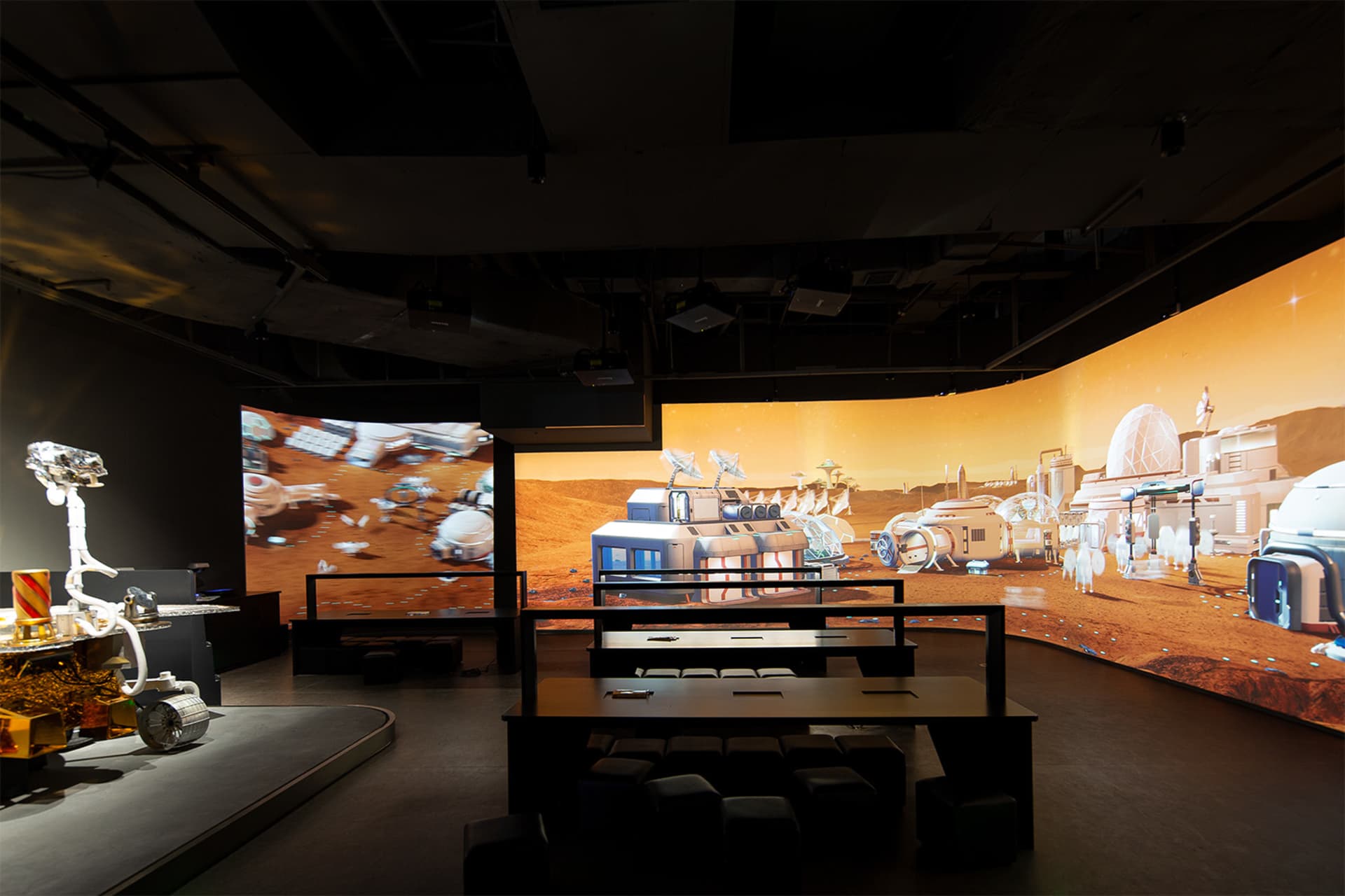 Interior view of a dark immersive exhibition hall with black modular seating, a large curved LED screen showing a Mars colony, and a white Mars rover model on the left.