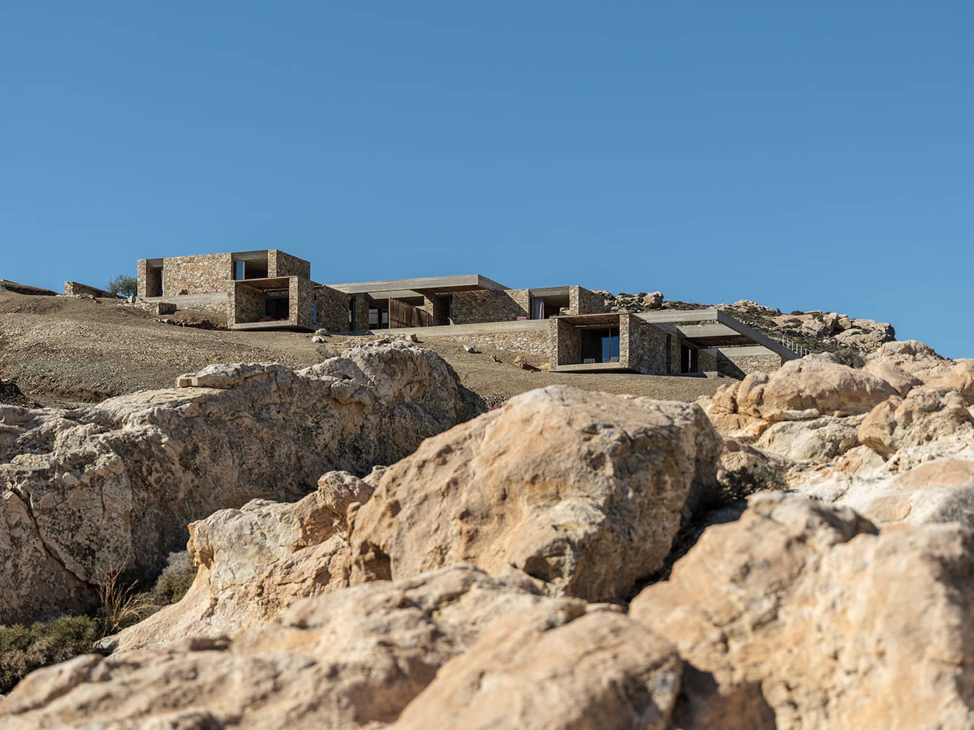 Modern cliffside villa constructed with local limestone blocks and concrete cantilevers, perched on rocky terrain with dramatic sea views under a clear blue sky.