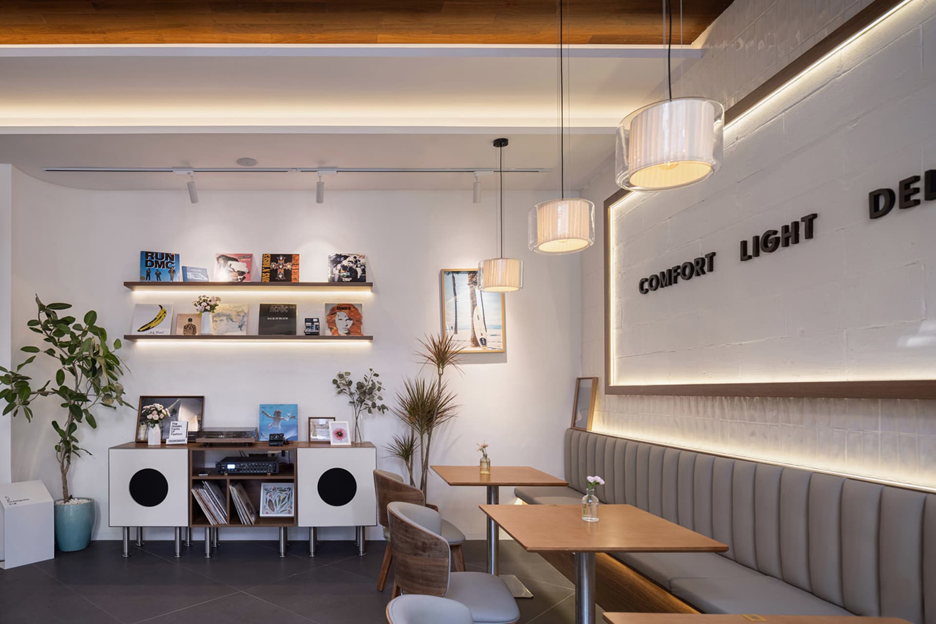 Modern cafe interior with wooden dining tables, grey upholstered banquette seating, white brick wall with backlit signage, floating wooden shelves displaying vinyl records, and warm pendant lighting.