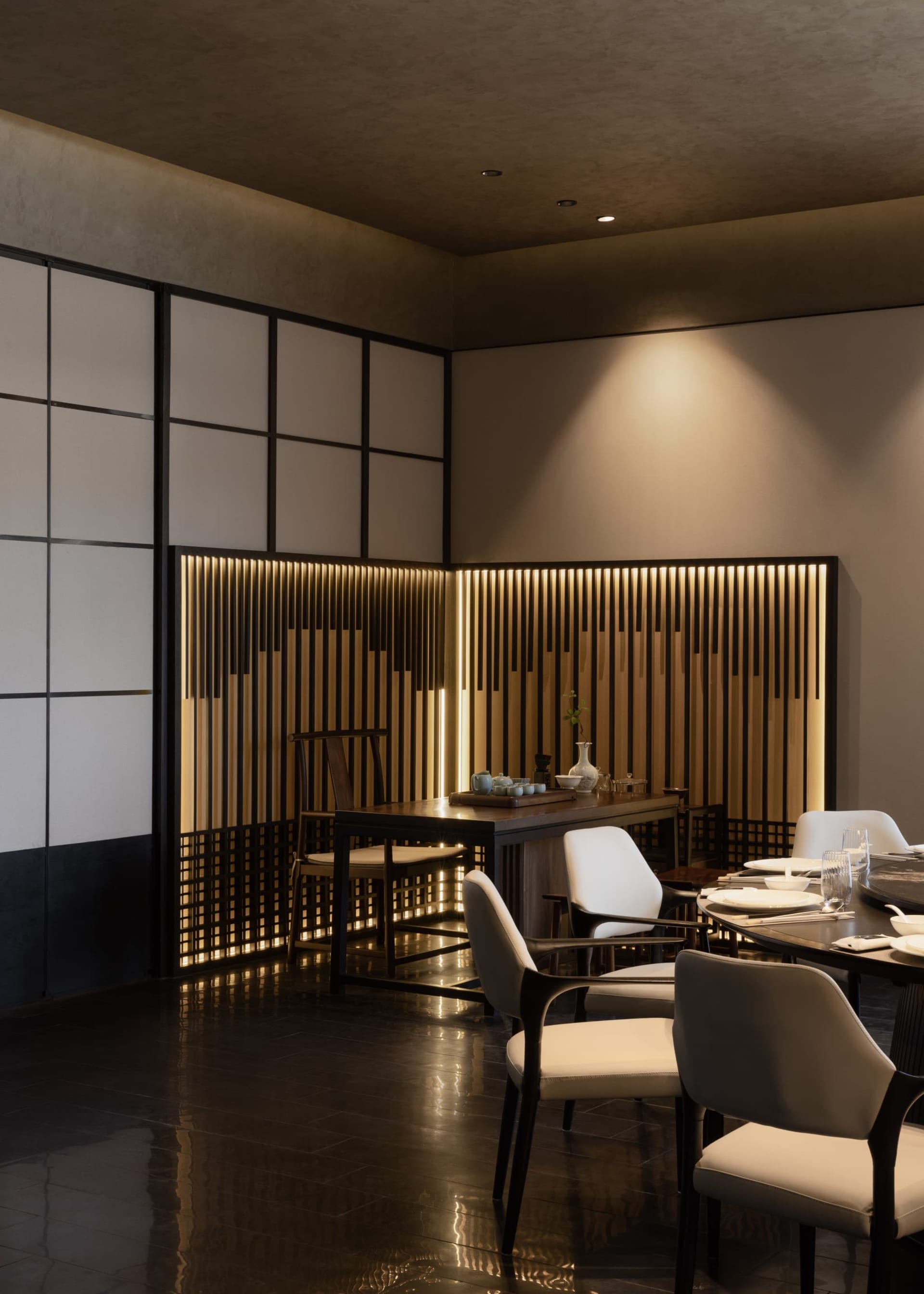 Modern dining room featuring a dramatic illuminated walnut slat wall, dark wood dining table, and cream upholstered chairs on polished dark tile flooring.