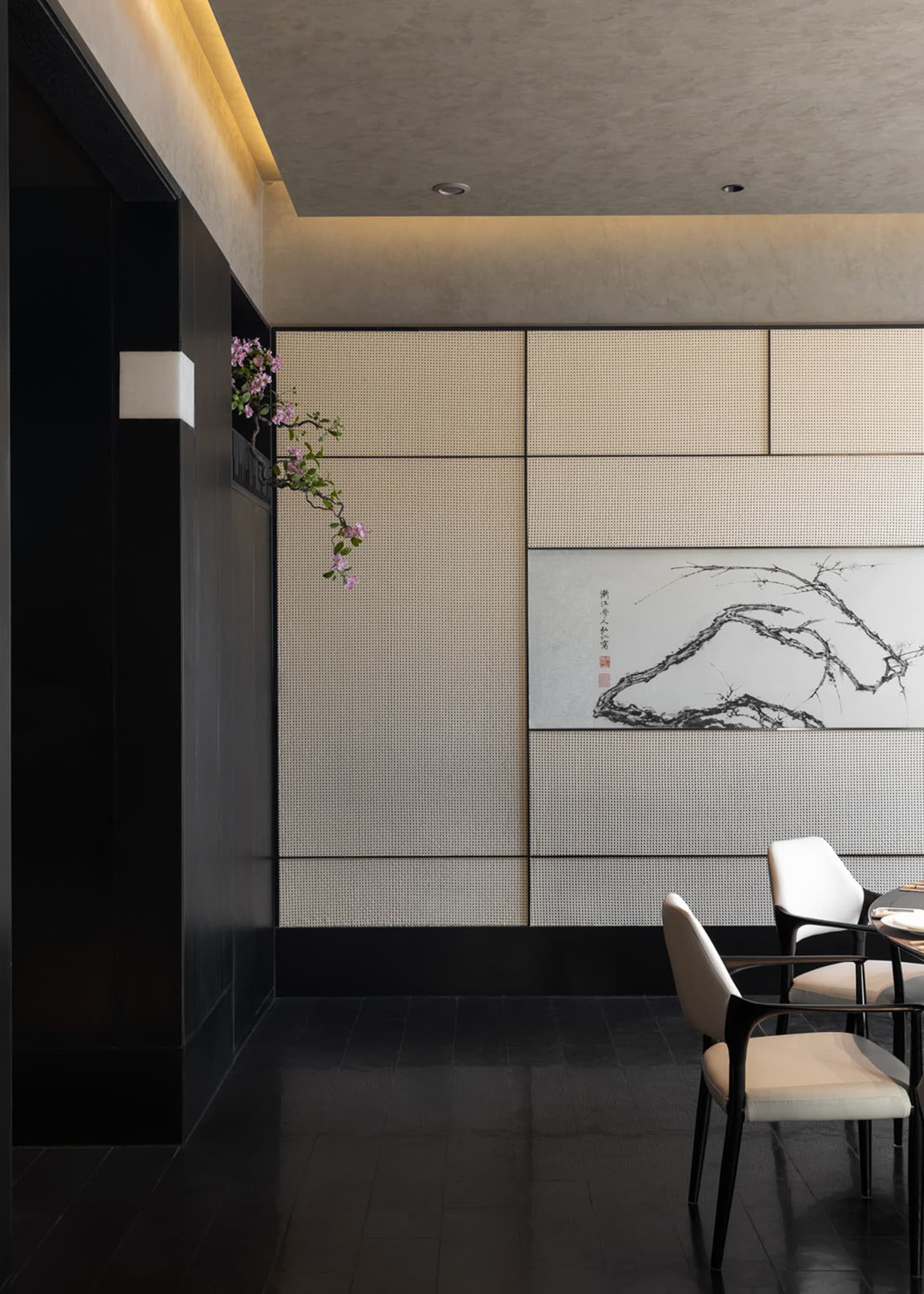 Modern Chinese-style dining room featuring dark wood furniture, cream textured wall panels, a large ink wash painting of a mountain and tree branch, and a hanging pink flowering branch.