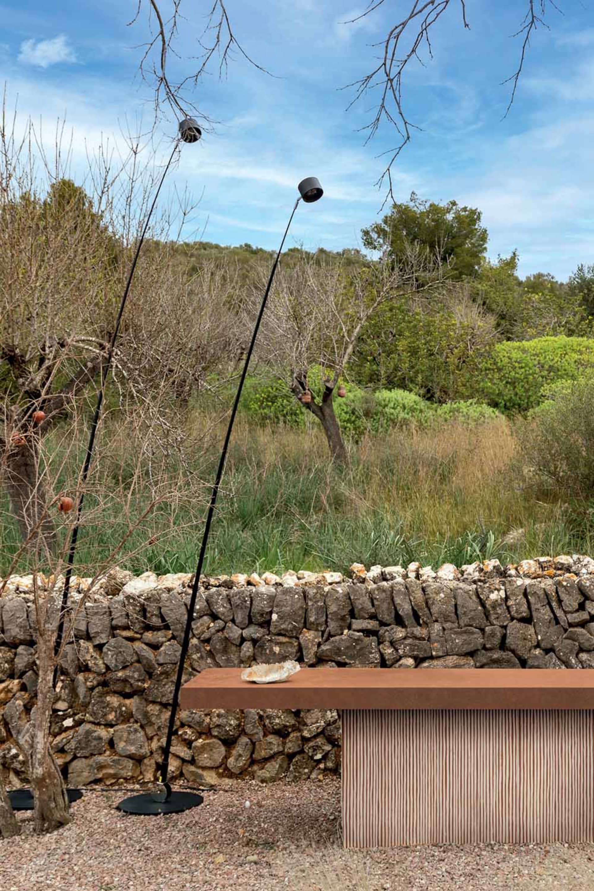 A modern terracotta console table with vertical ribbing placed against a rustic dry-stone wall in a Mediterranean garden with bare fruit trees and black sculptural floor lamps under a blue sky.