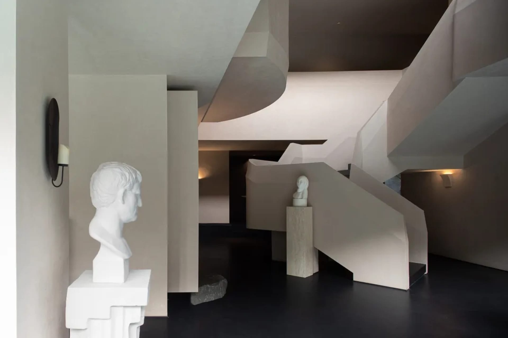 Modern interior featuring an angular concrete staircase, white classical marble busts on pedestals, and minimalist lighting.