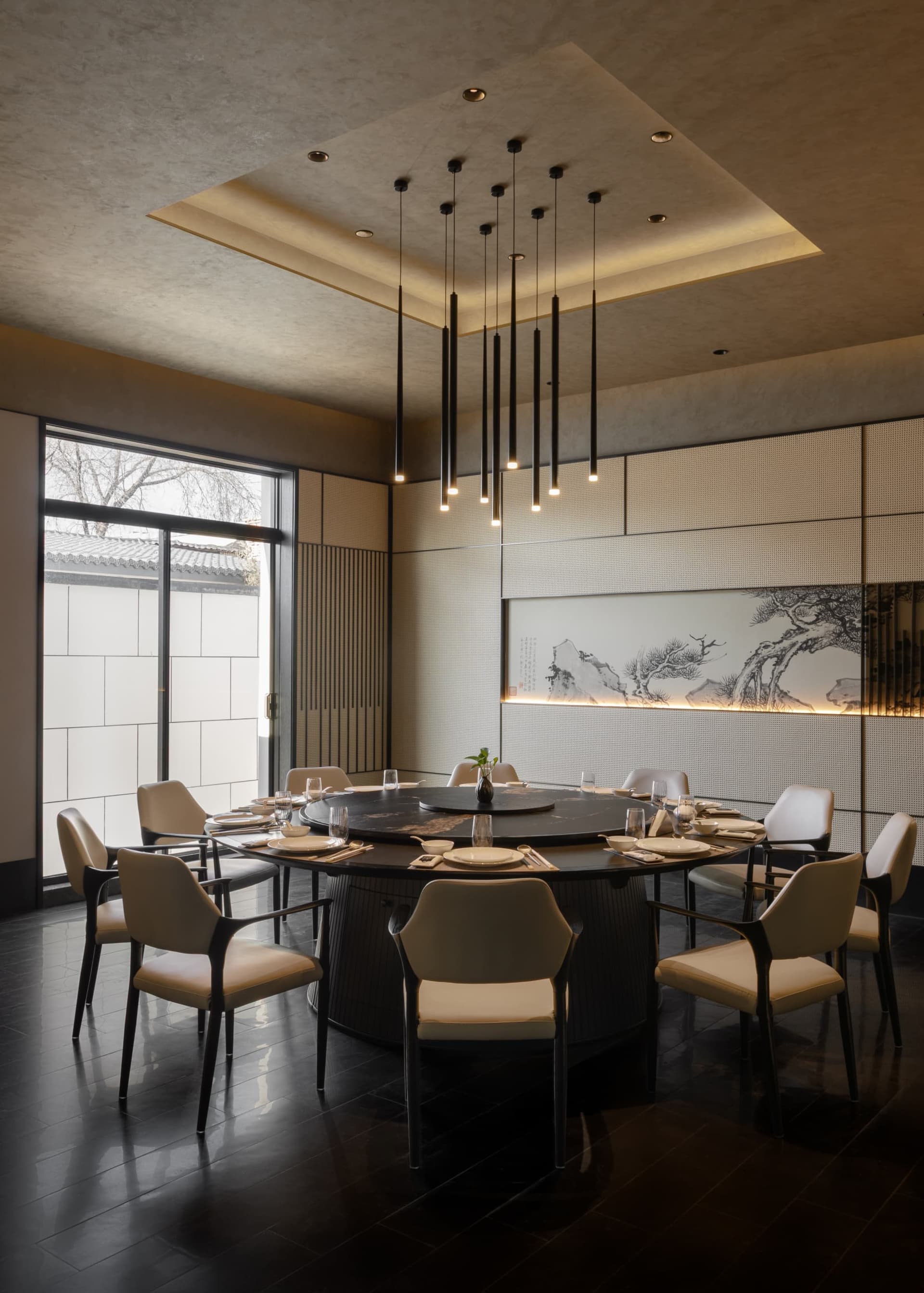 Elegant modern Chinese-style dining room featuring a large black marble round table, ten cream upholstered chairs, dramatic black pendant lights, and a backlit Chinese ink painting of pine trees and mountains.