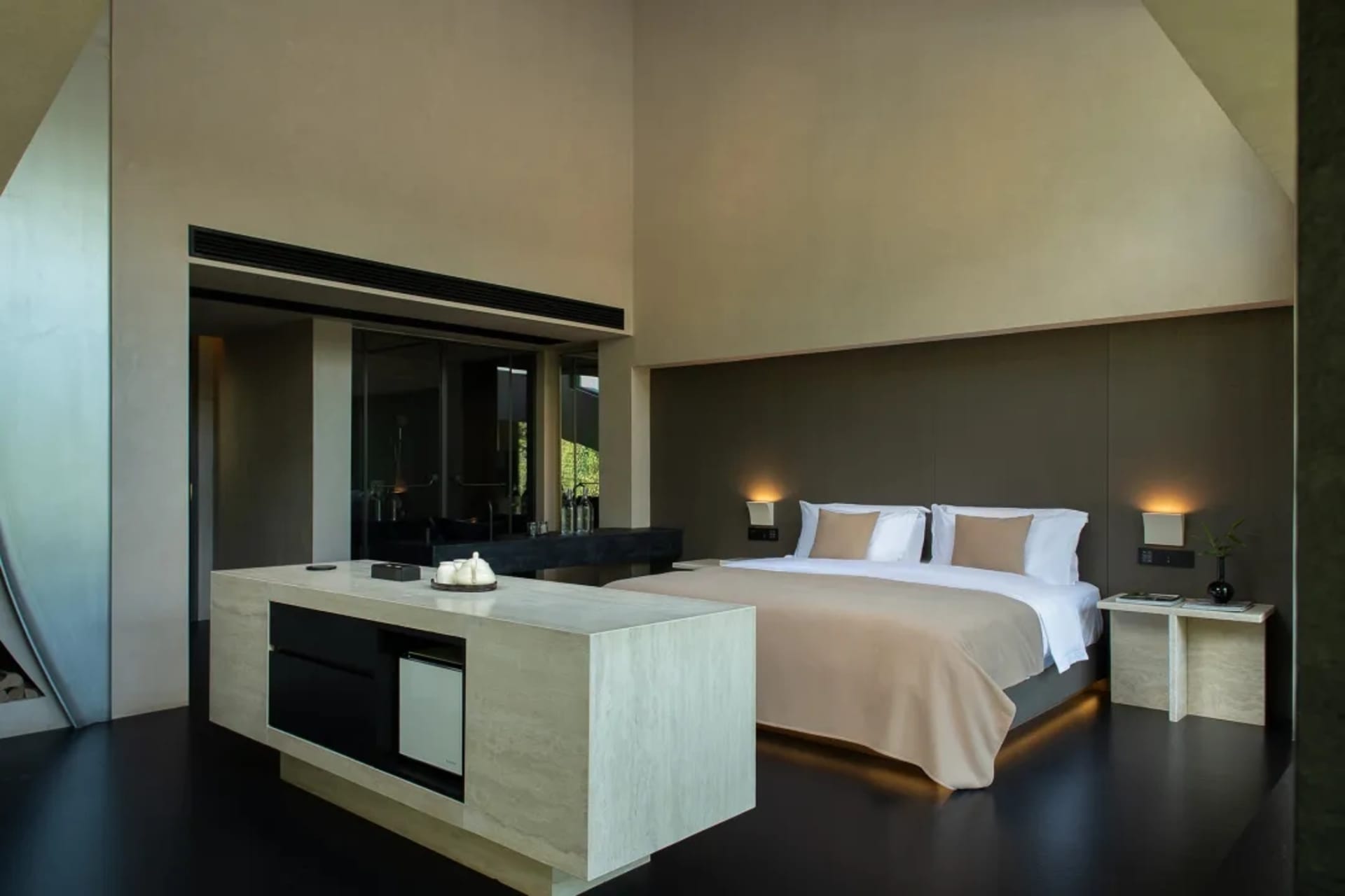 Modern minimalist bedroom featuring a large travertine stone island, low platform bed with beige bedding, dark polished floor, and warm wall sconces under high angled ceilings.