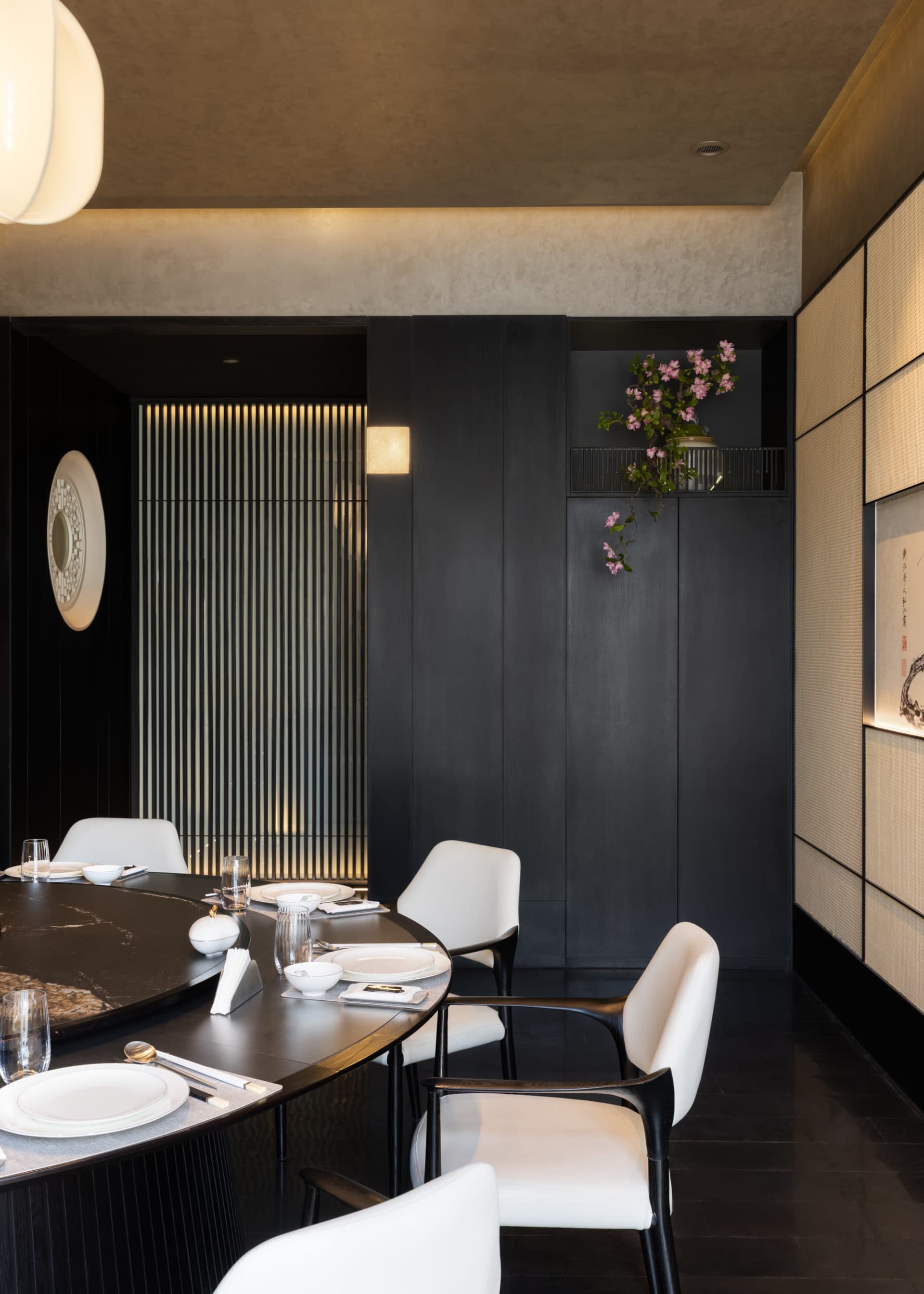 Modern dark-toned dining room featuring a large round black marble table, white upholstered dining chairs with dark wood frames, textured beige wall panels, vertical backlit slat wall feature, and pink cherry blossom arrangement in a built-in niche.