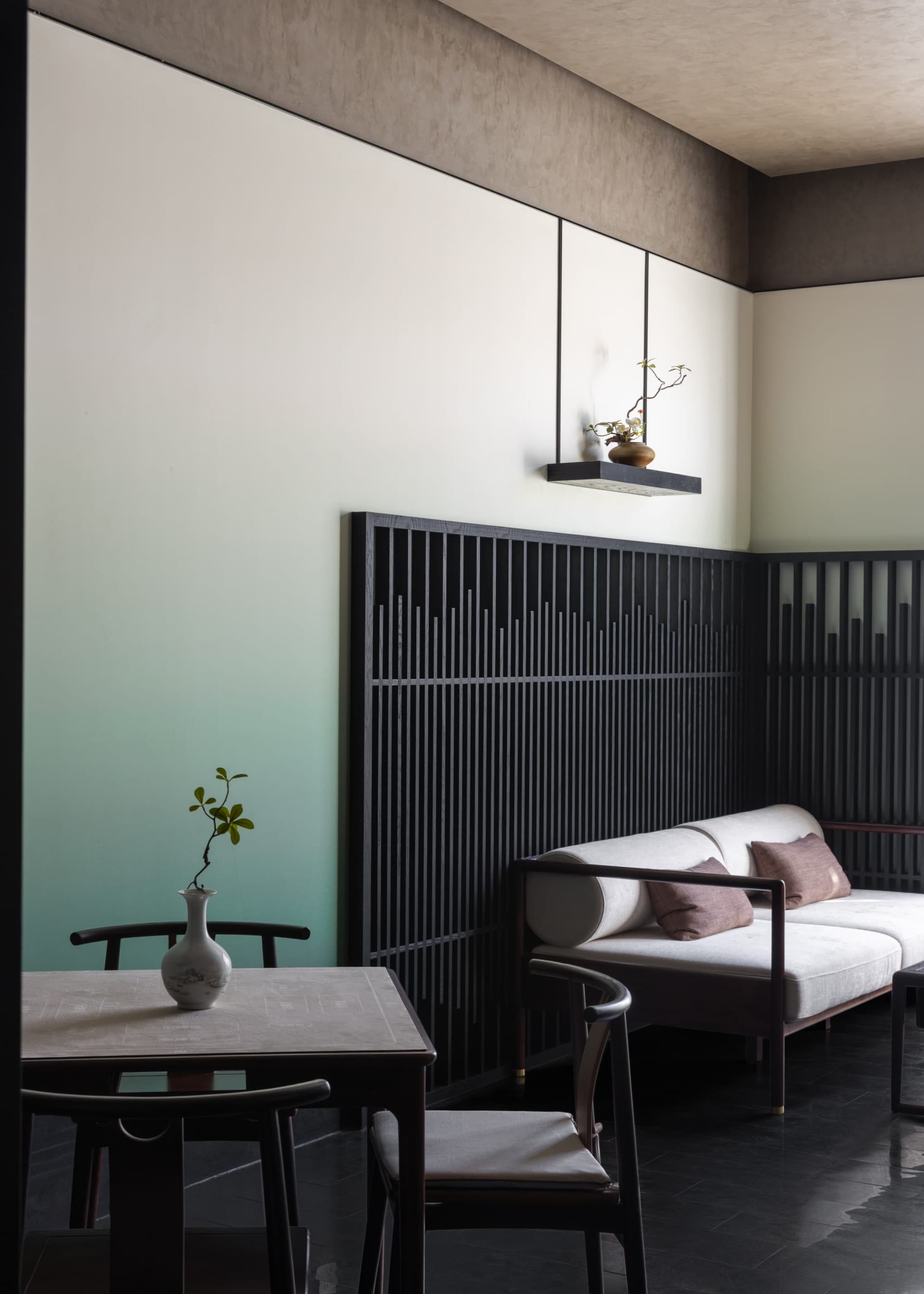 Modern Chinese-style living room featuring a walnut daybed with light upholstery, dark vertical slatted screen, muted green accent wall, and dark wood dining set with ceramic vase.