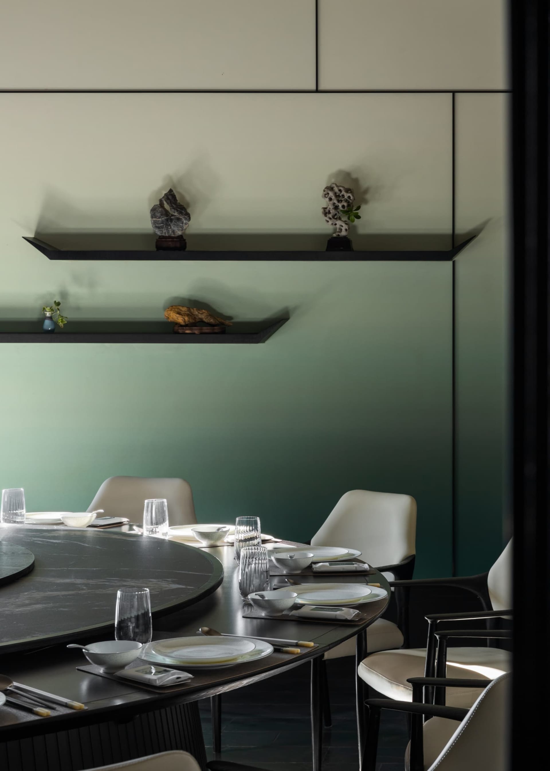 Modern dining room with round black marble table, white leather dining chairs, and floating shelves displaying decorative stones against a sage green gradient wall.