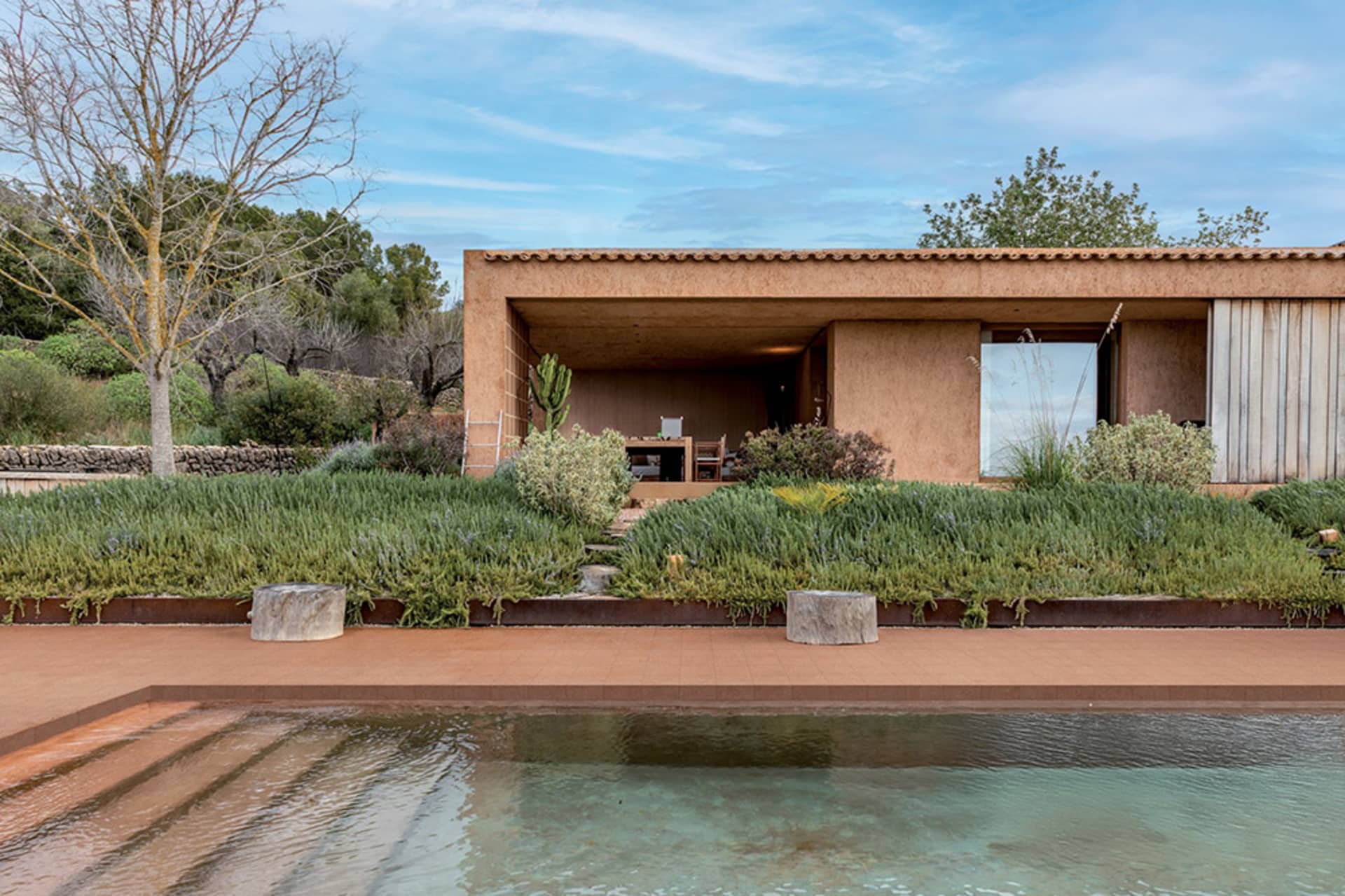 Modern terracotta villa with large glass openings, wooden elements, and a lush garden terrace beside a clear swimming pool under a blue sky.