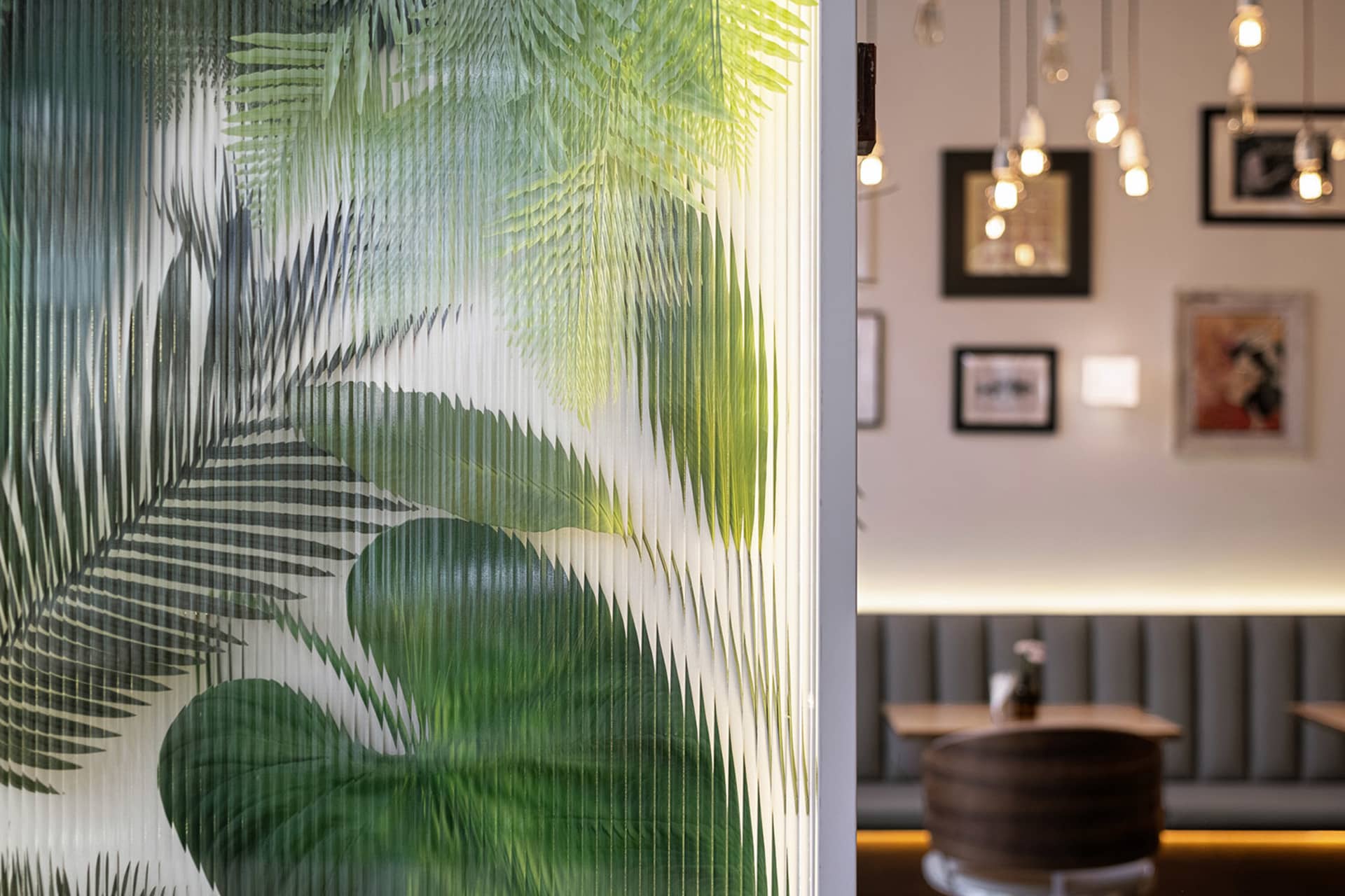 Close-up of a backlit fluted glass partition featuring large tropical palm and monstera leaf patterns in shades of green, with a modern restaurant seating area visible in the background.