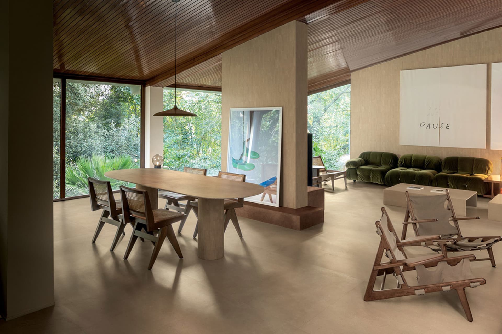 Modern open-plan living and dining area featuring a large wooden dining table with woven chairs, travertine flooring, wooden ceiling, green velvet sofa, and large windows overlooking a lush forest garden.