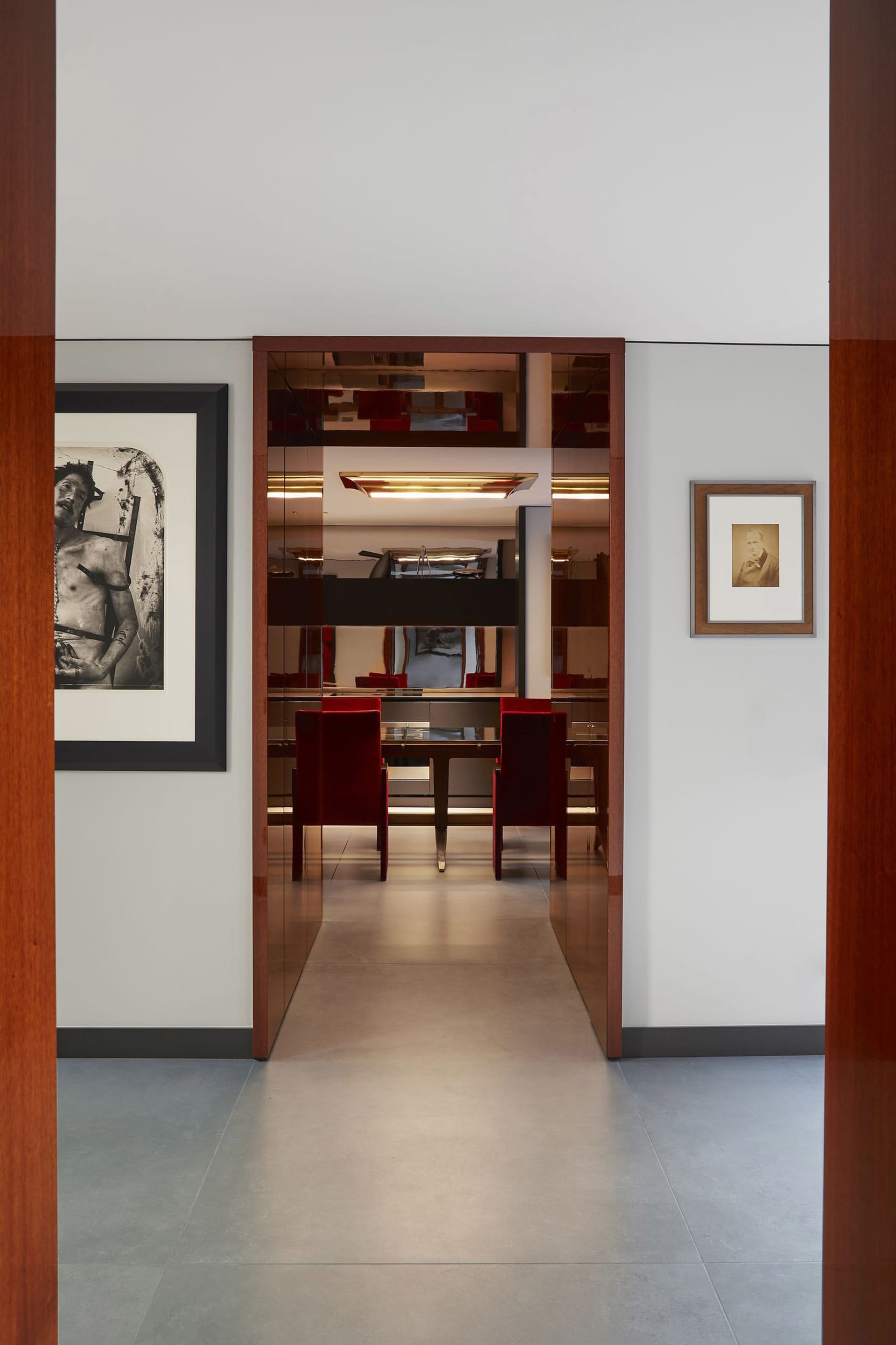 View through a doorway into a modern dining room featuring mirrored walls, red velvet dining chairs, dark wood accents, and contemporary art on white walls.