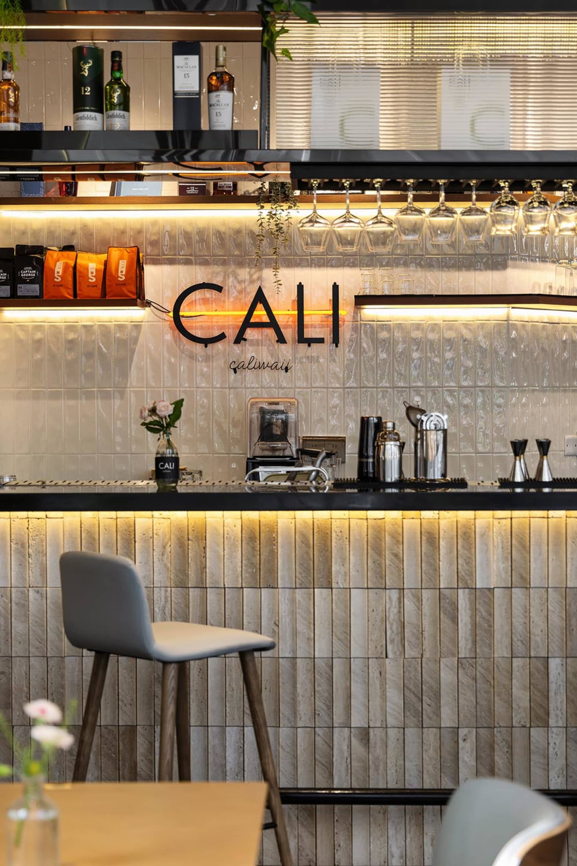 Modern cafe bar interior with grey textured tile wall, black countertop, grey bar stool, hanging wine glasses, and warm LED strip lighting.