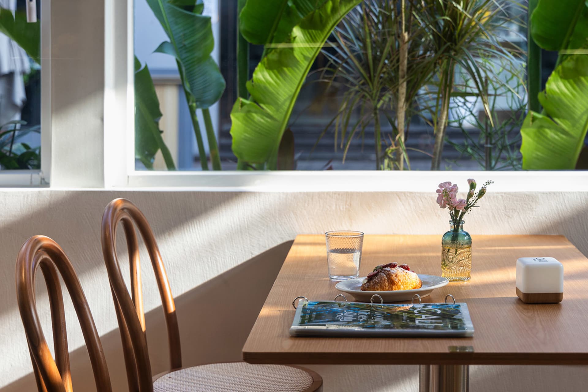 Wooden cafe table by a large window with green plants outside, featuring a croissant on a plate, glass of water, small flower vase, and two bentwood chairs in warm sunlight.