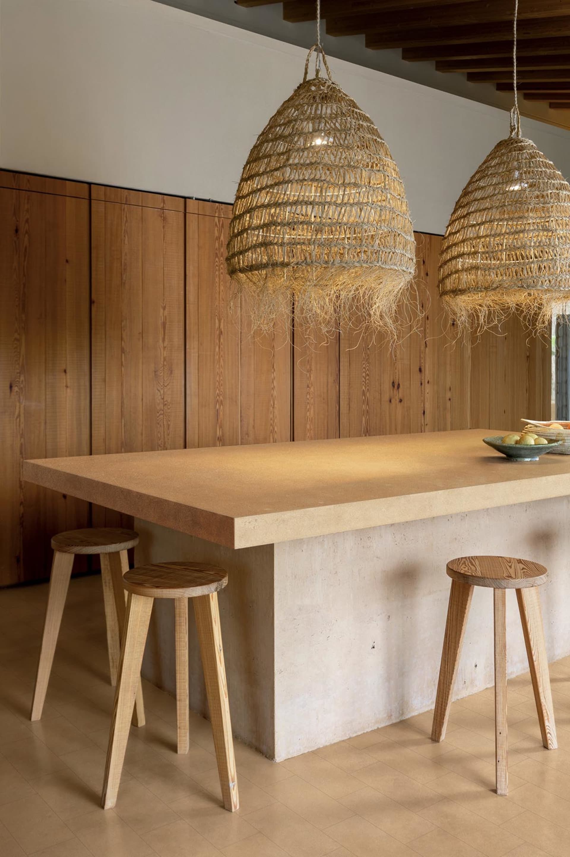 Modern minimalist kitchen island made of concrete and wood, paired with two large woven rattan pendant lights and three wooden stools against a vertical wood-paneled wall.