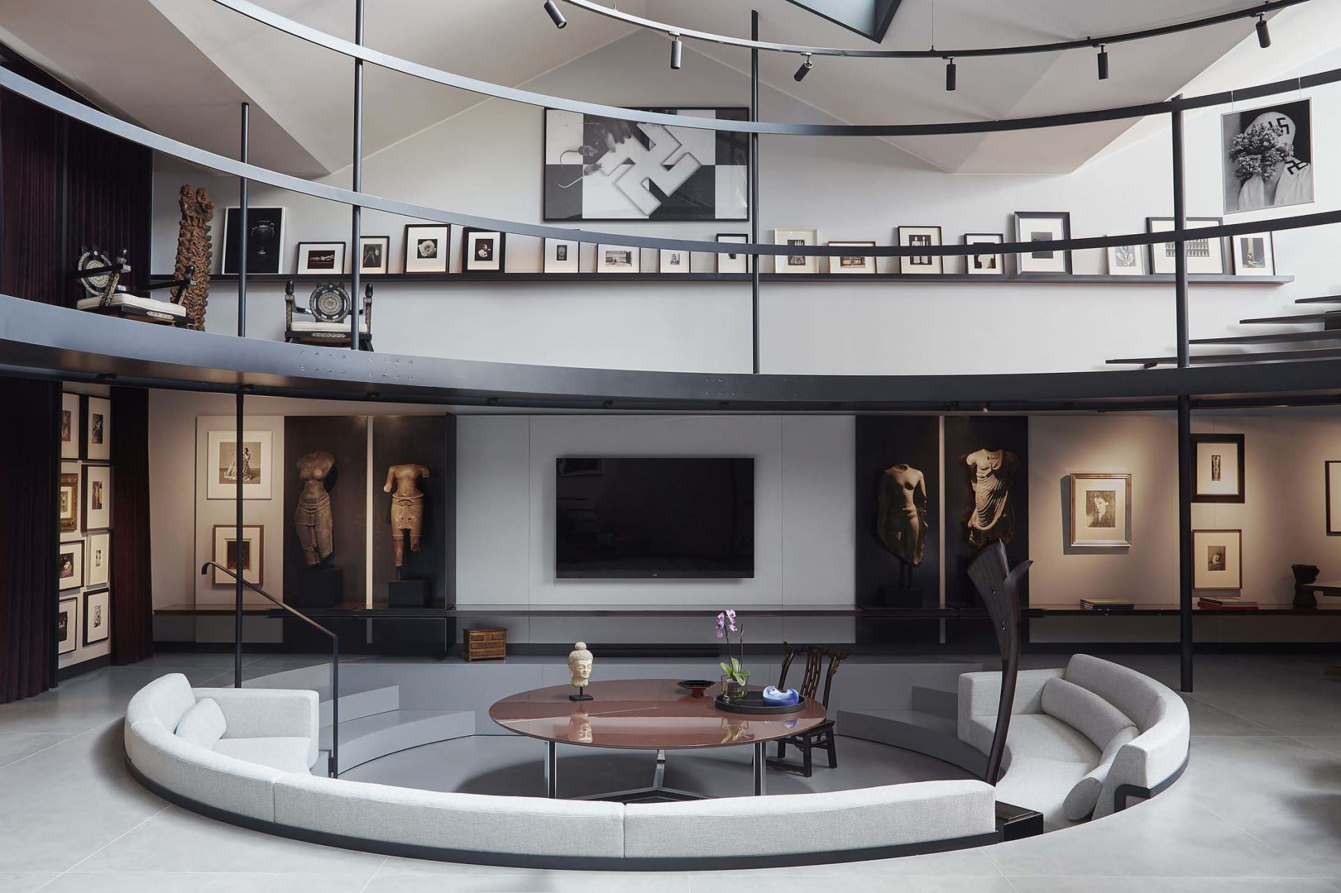 Modern double-height living room featuring a large curved light-grey sectional sofa, circular rosewood coffee table, extensive art collection on two levels, and classical stone sculptures displayed throughout the space.