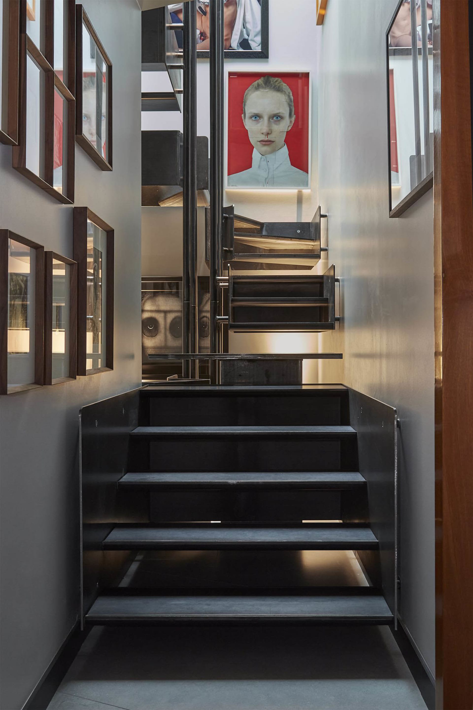 Modern black steel floating staircase with integrated lighting, flanked by dark wood framed portraits and a large red portrait of a woman at the top landing.