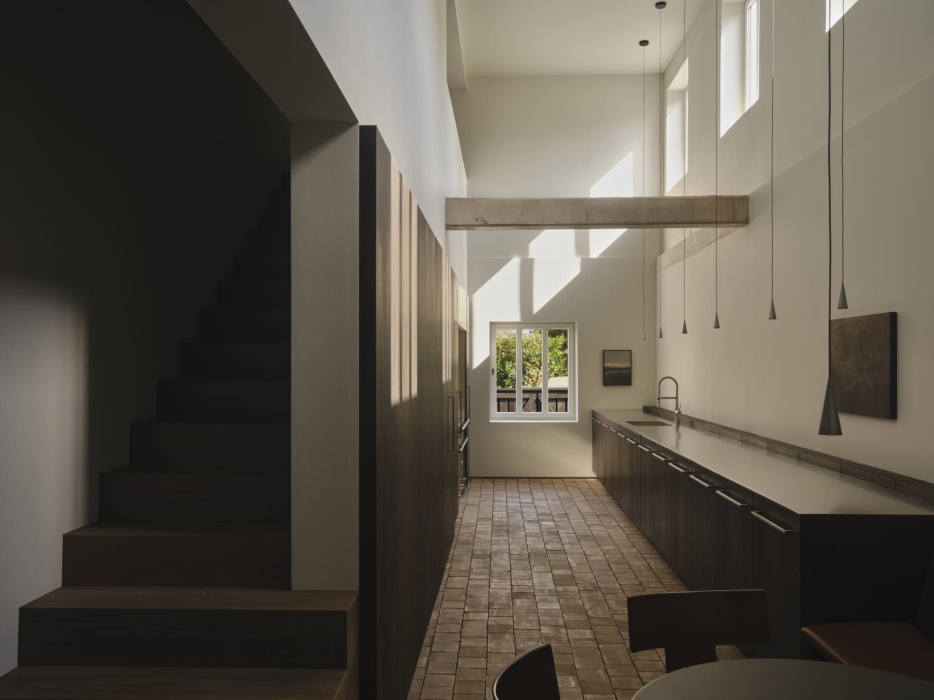 Modern minimalist kitchen and dining area with dark wood cabinetry, long white countertop, pendant lights, exposed concrete beam, and wooden staircase on the left.