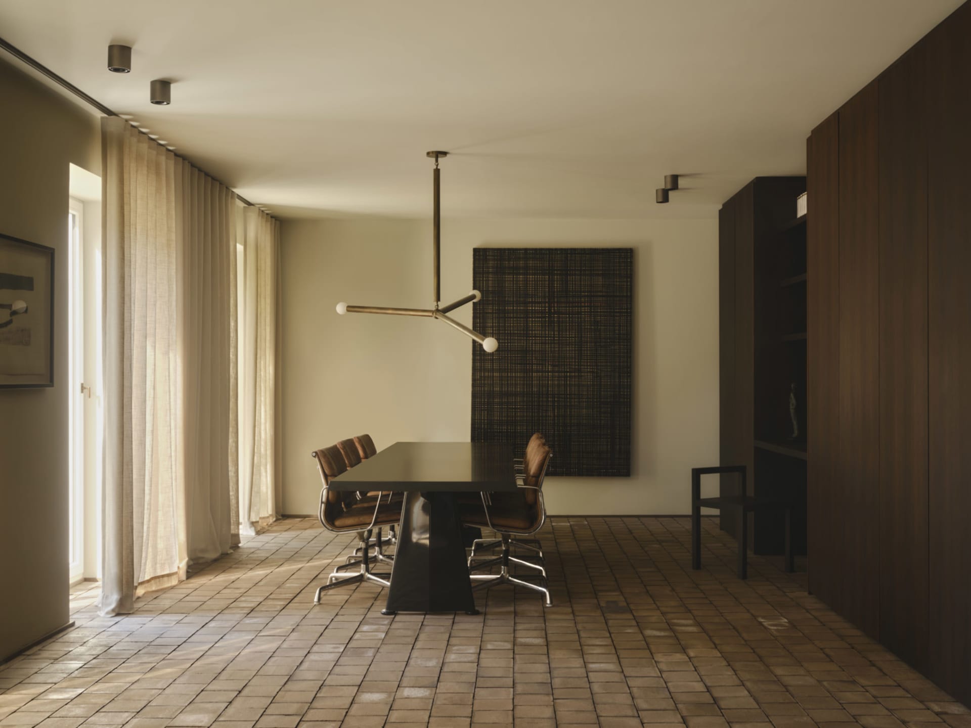 Modern minimalist dining room featuring a dark dining table, brown leather chairs, walnut wood paneling, large woven wall art, and light terrazzo tile flooring with sheer curtains allowing natural light.