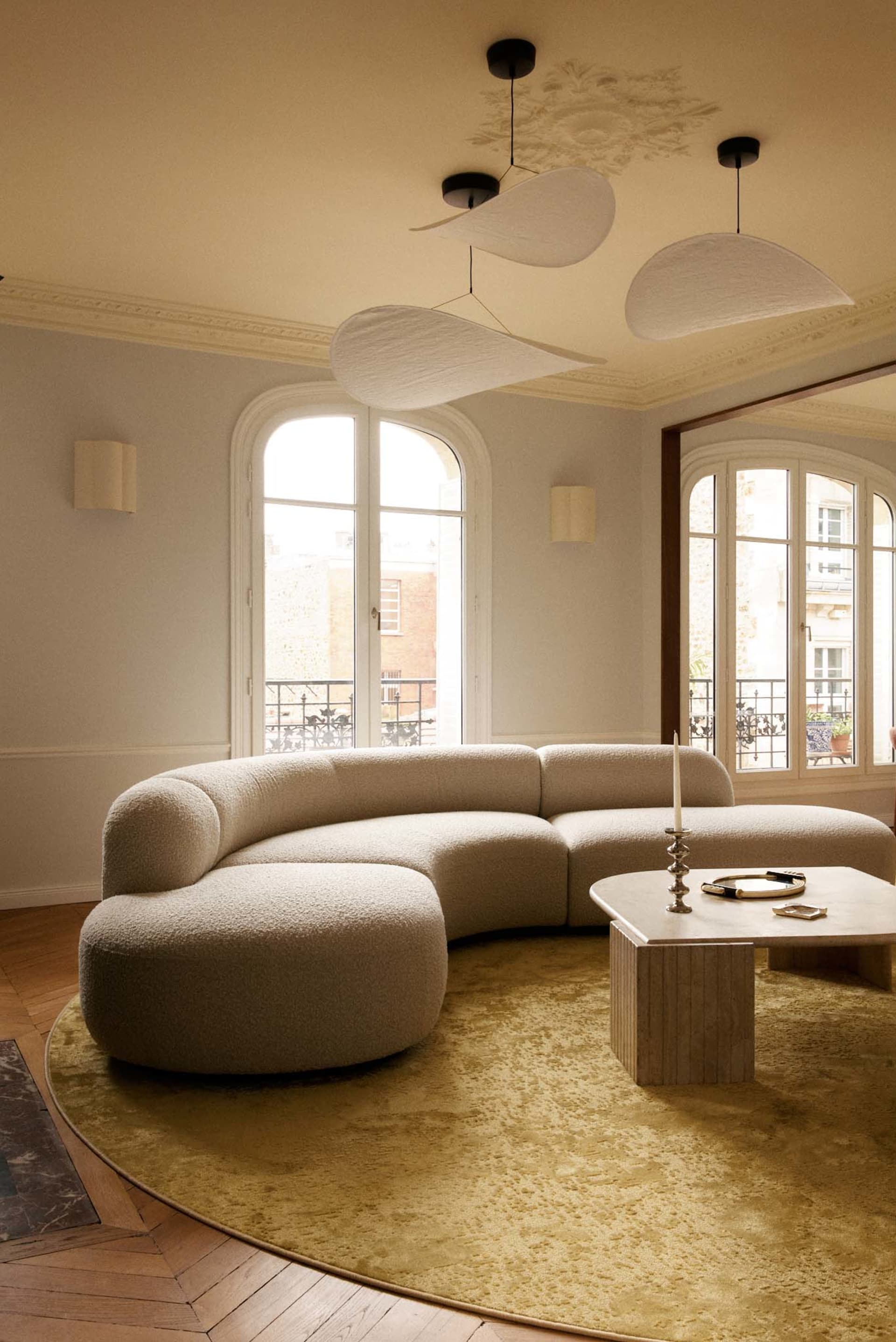 A spacious Parisian living room featuring a large curved off-white boucle sectional sofa on a round chartreuse rug, with three sculptural white pendant lights hanging from a ceiling with ornate plasterwork, arched French windows, and herringbone wood flooring.
