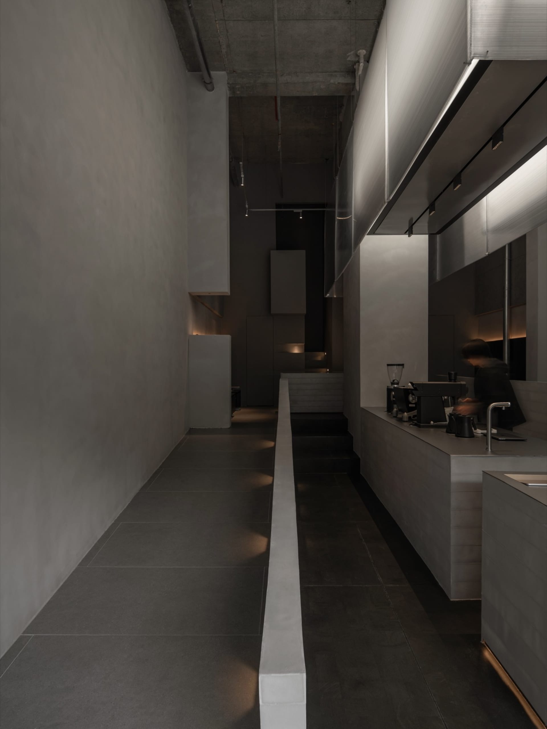 Long narrow corridor in a brutalist-style commercial kitchen with raw concrete walls, dark tiled floors, linear LED lighting, and a person operating an espresso machine on the right.