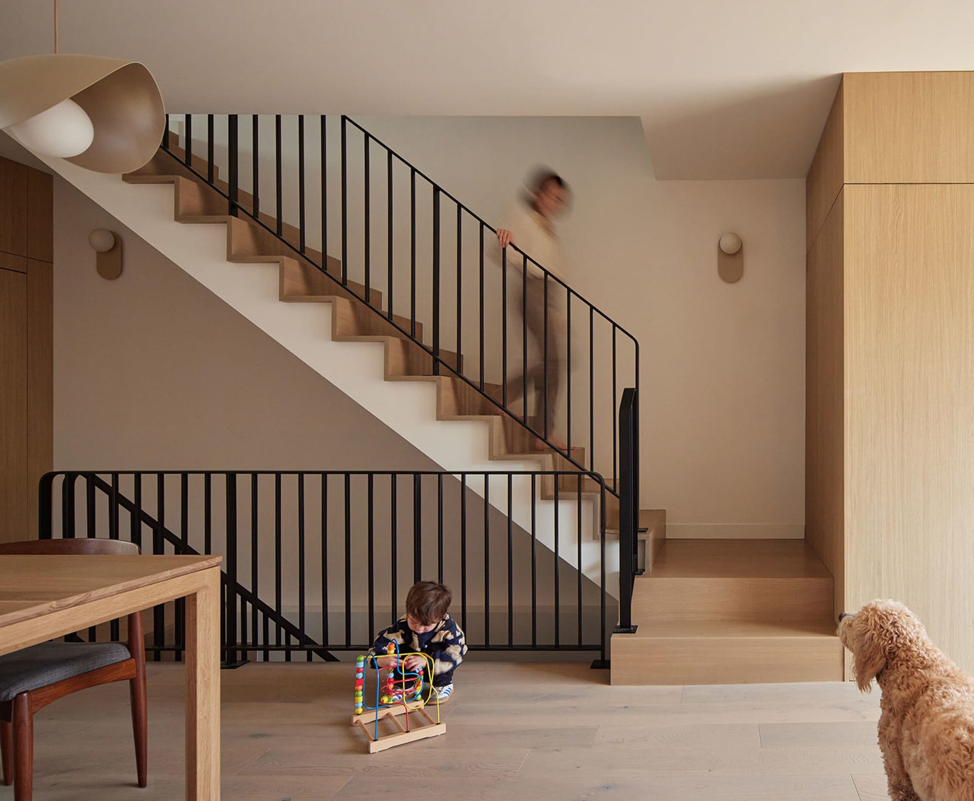 Modern family home interior featuring a light oak staircase with black metal railings, wooden dining table, pendant light, child playing, and a dog in the foreground.