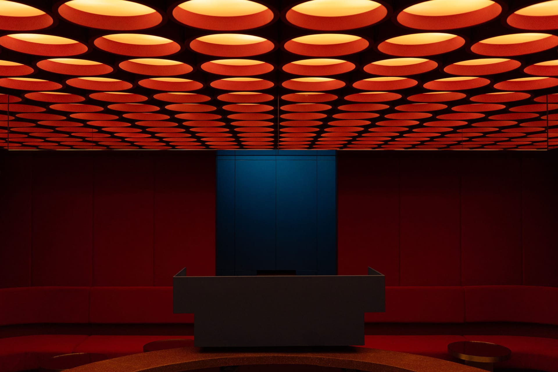 Modern theater auditorium featuring deep red velvet seating, a dark stage backdrop, and a ceiling covered in glowing orange circular light fixtures.