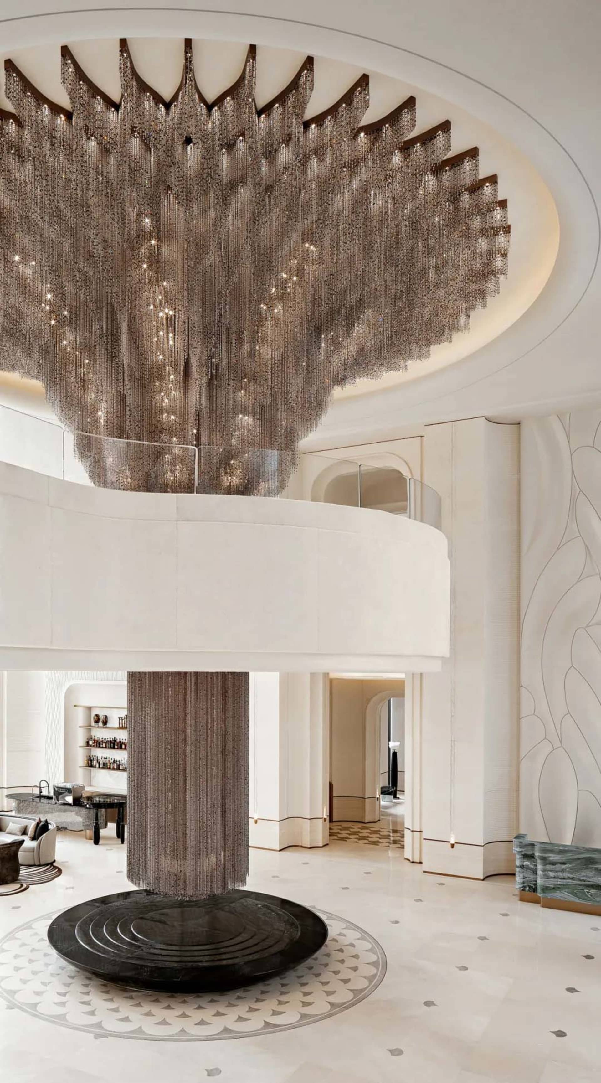 Expansive luxury hotel lobby featuring an enormous cascading crystal-bead chandelier hanging above a circular black marble base, with a curved mezzanine and light stone finishes.