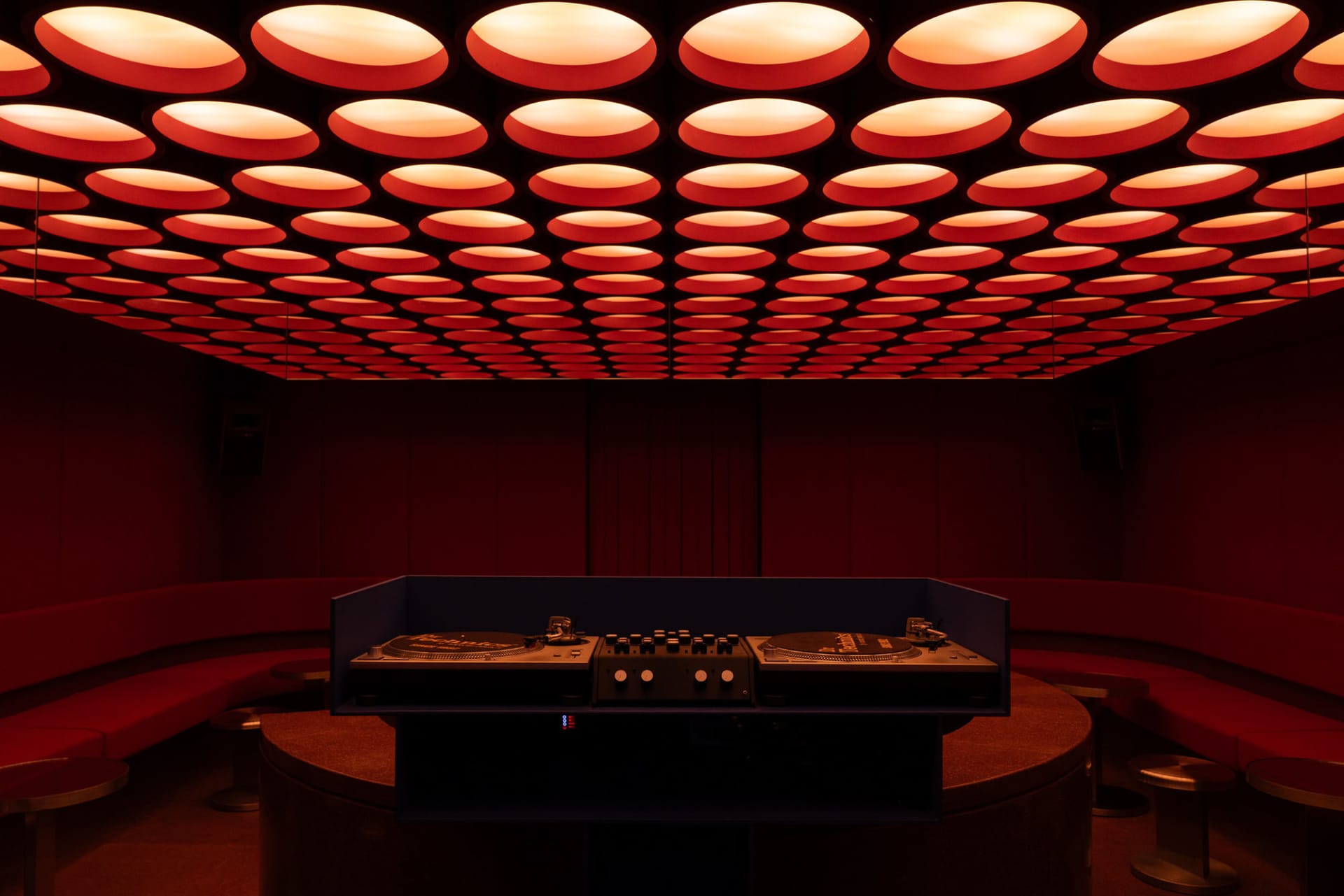 Interior view of a nightclub DJ booth featuring two turntables and mixer on a curved console, surrounded by red velvet curved banquettes under a ceiling covered with dozens of glowing red circular light panels.