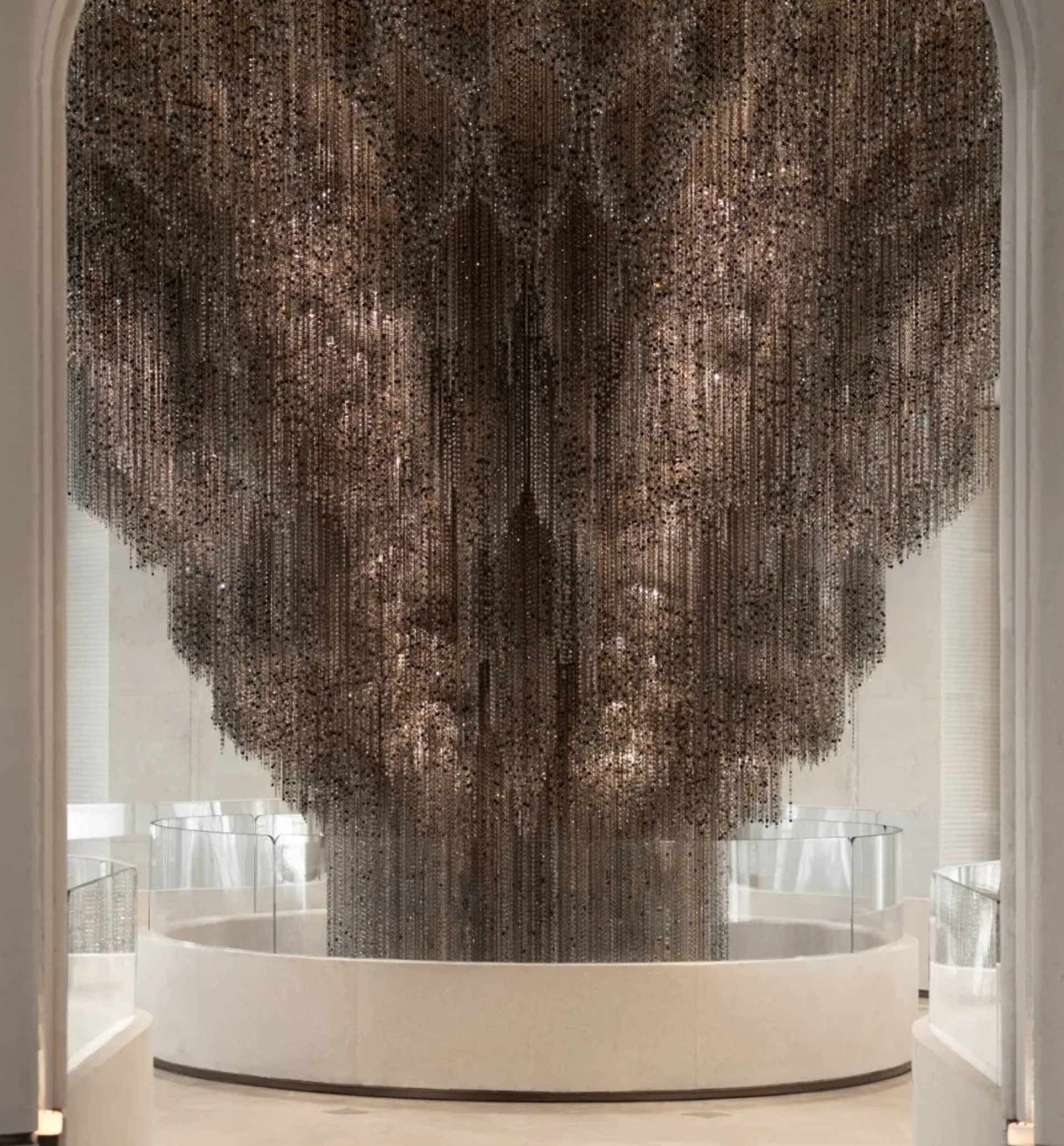 Massive sculptural art installation composed of cascading dark crystal beads hanging from a high ceiling in a modern grand lobby with white marble and glass railings.