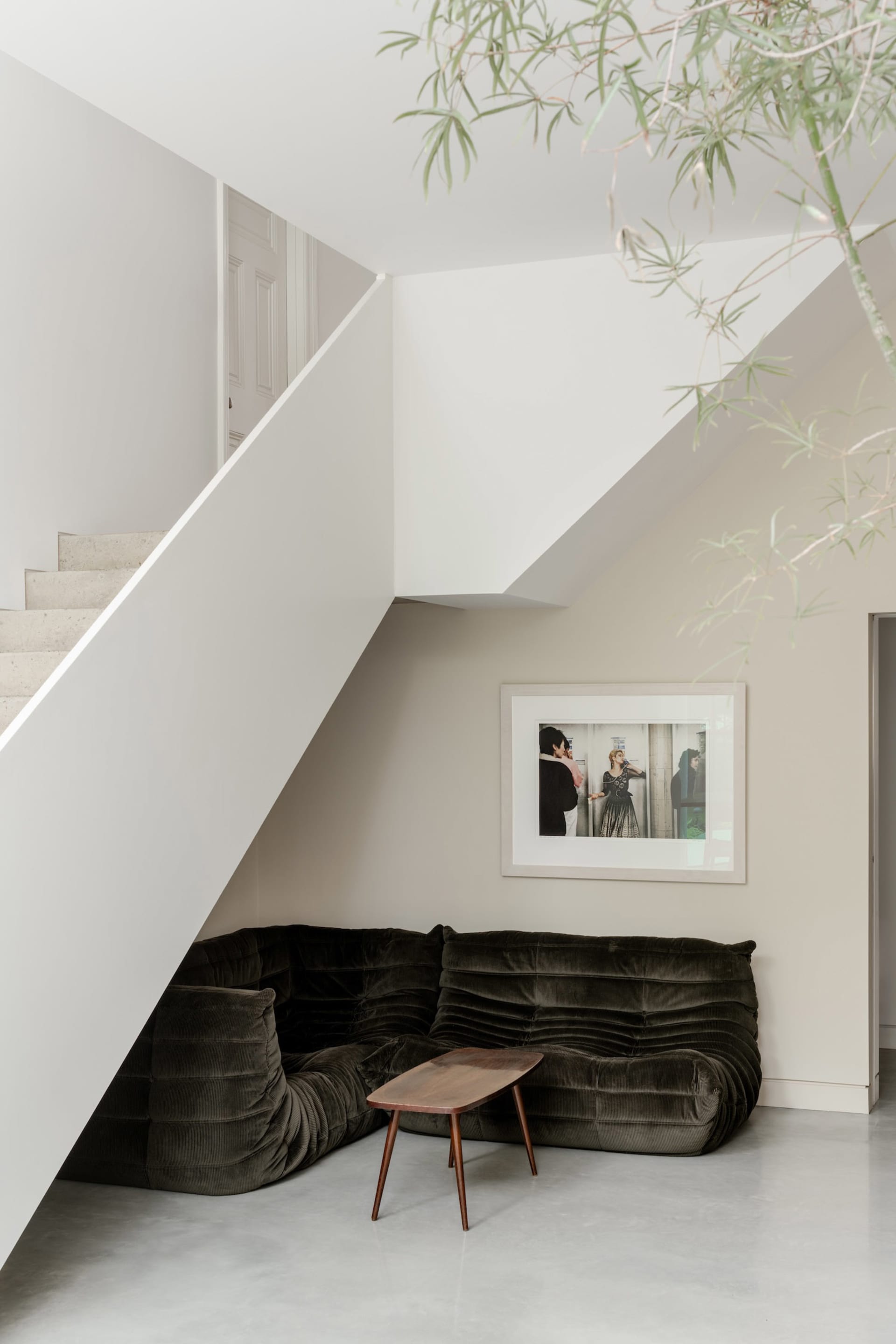 Modern minimalist living space featuring a dark green corduroy modular sofa under a white staircase, accompanied by a walnut coffee table and framed black and white photograph on the wall.