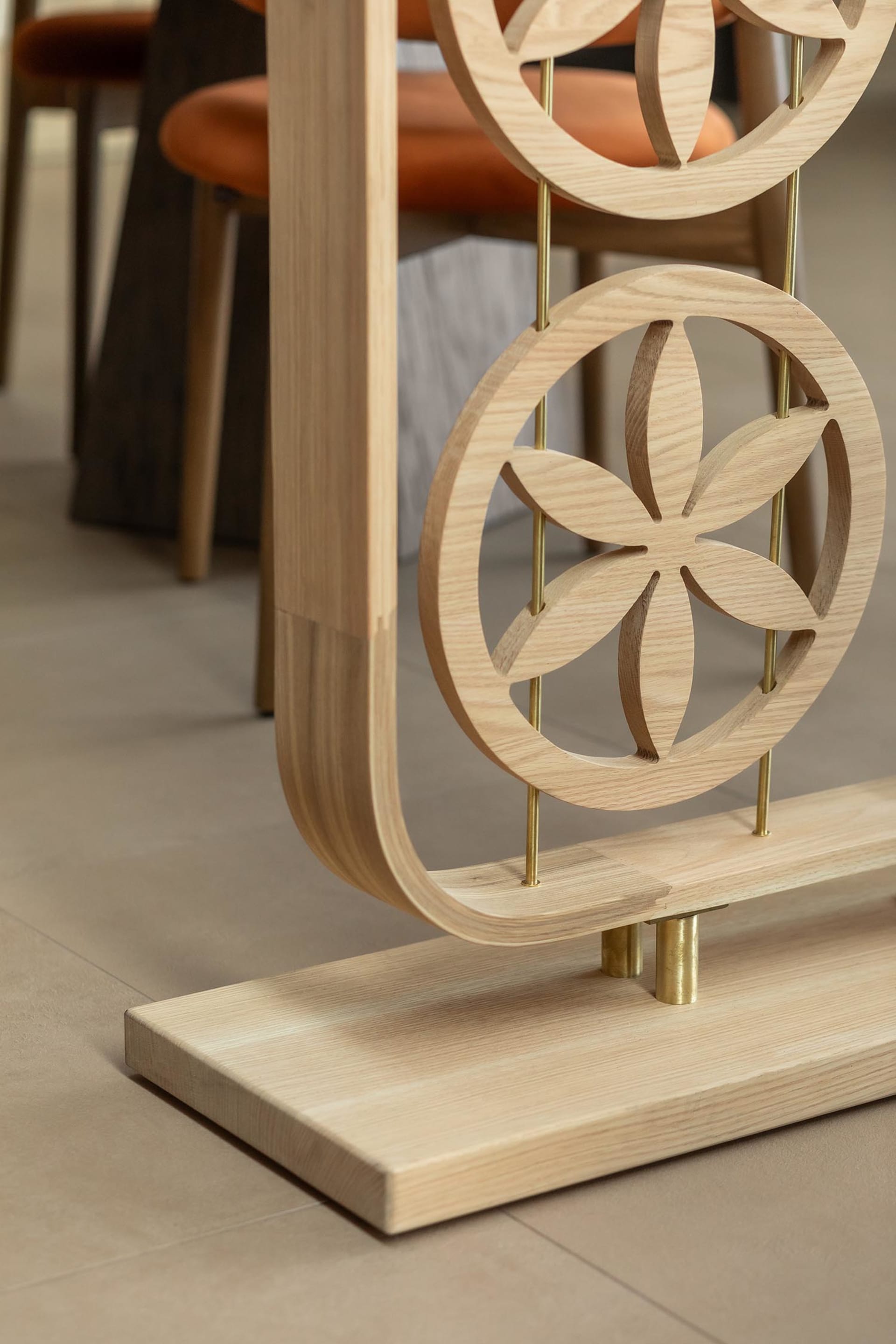 Close-up of a light oak room divider with circular floral cutout patterns, brass rods, and a curved wooden base, with orange dining chairs visible in the background.