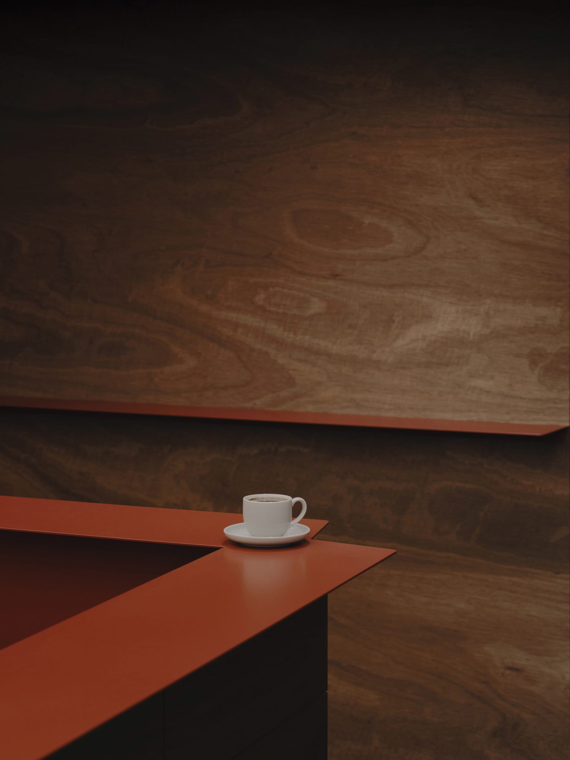 Close-up of a white espresso cup and saucer on a red counter with warm walnut wood wall in the background